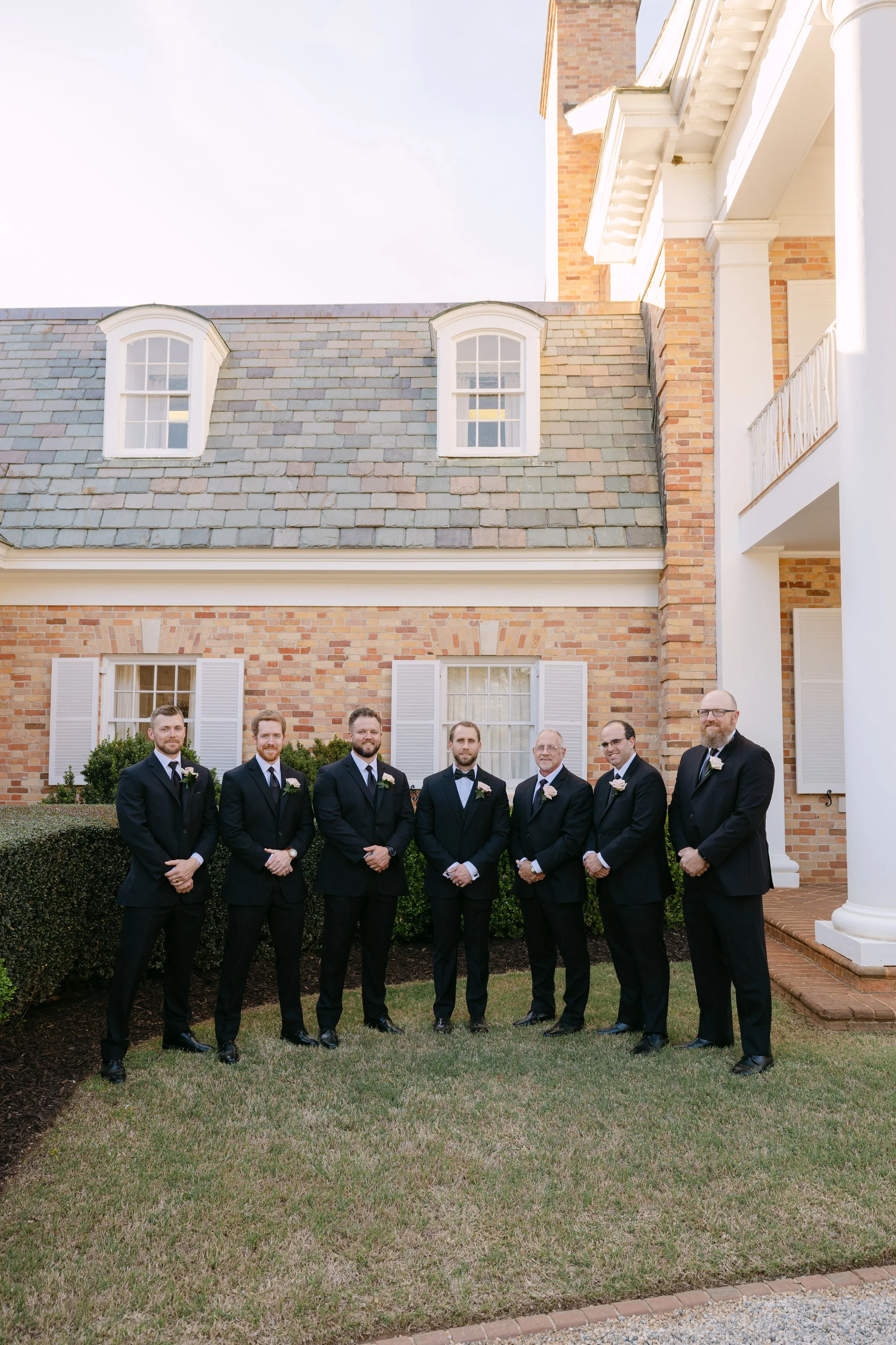 Group of six men in black tuxedos standing on grass in front of a brick building with white shutters and a columned porch.