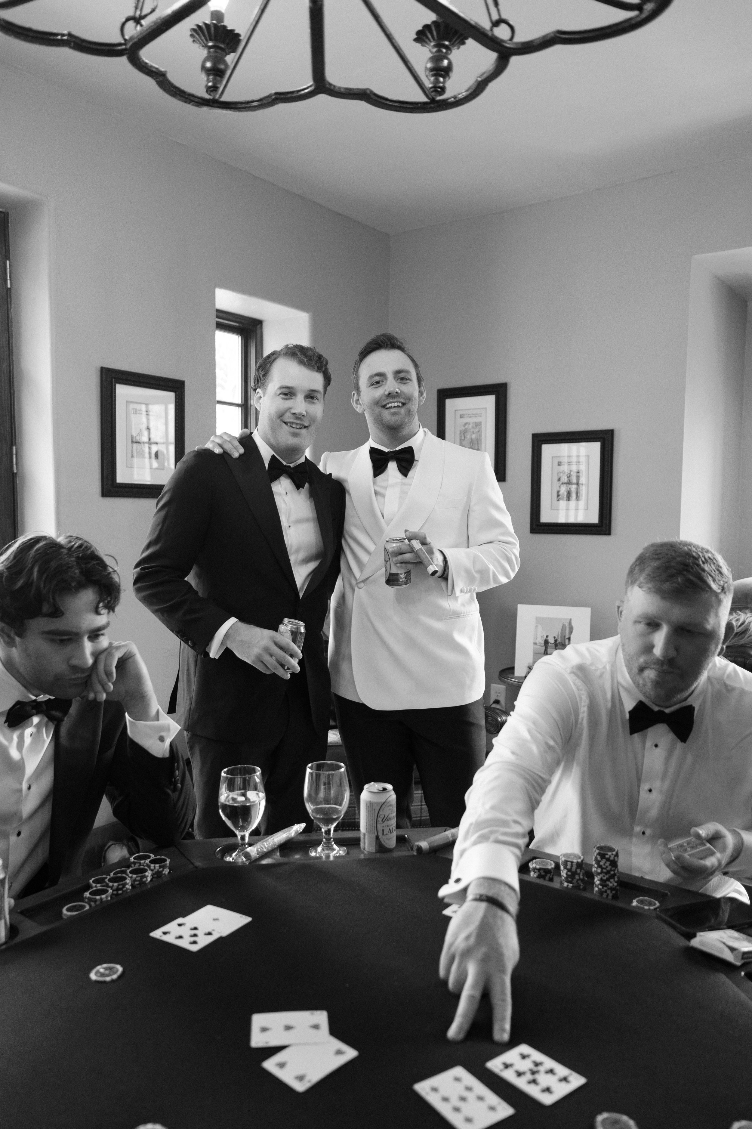 Black and white photo of five men in tuxedos, two standing and three seated at a poker table, playing cards and chips, in a well-lit room with framed pictures on the walls.