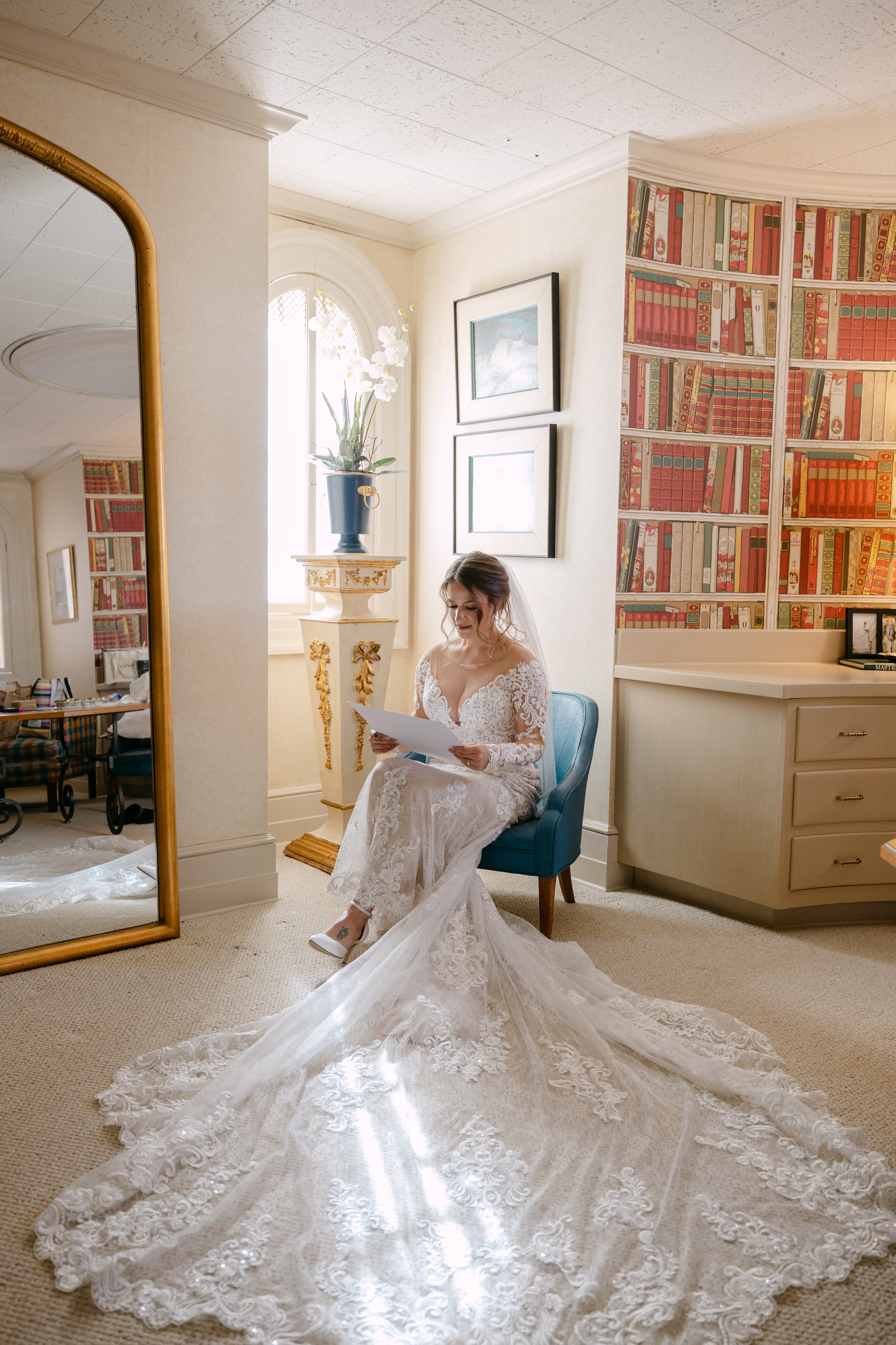 Bridal bride sitting on blue chair in a hotel room, wearing a lace wedding dress with a long train, reading a note or letter, with a large mirror and bookshelf in the background.