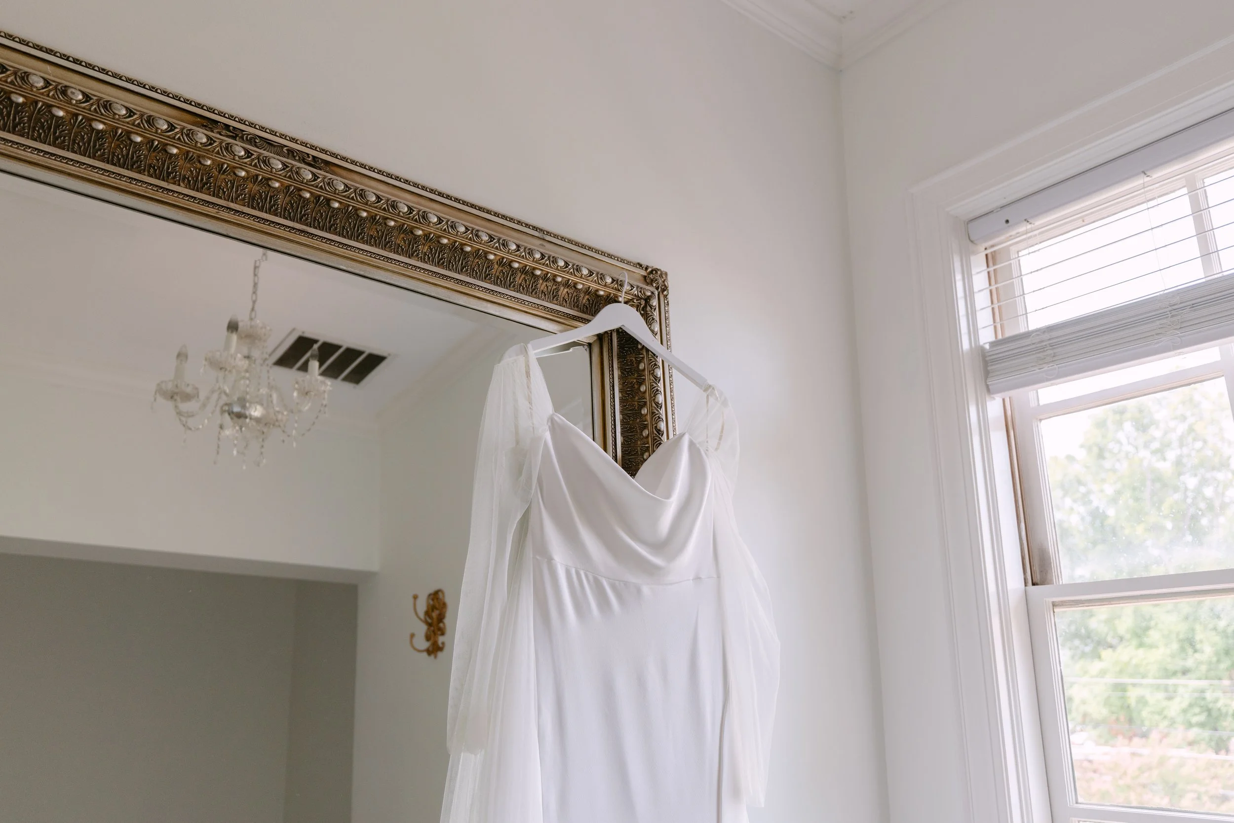 A white wedding gown hanging on a white hanger in front of an ornate gold mirror in a bright room with a chandelier and a window with white blinds.