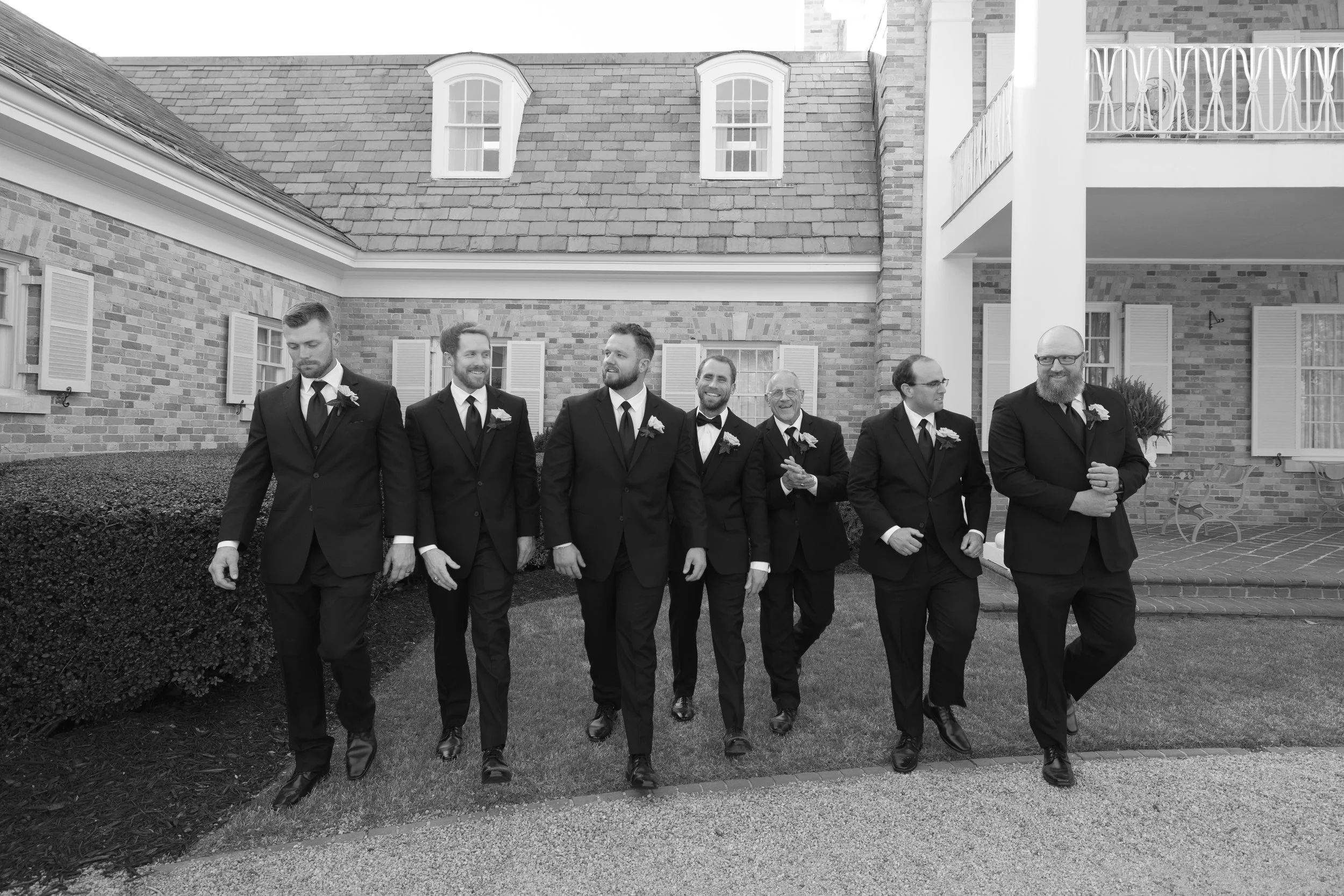 A group of seven men in tuxedos walking together outside a brick house with shutters and a balcony.
