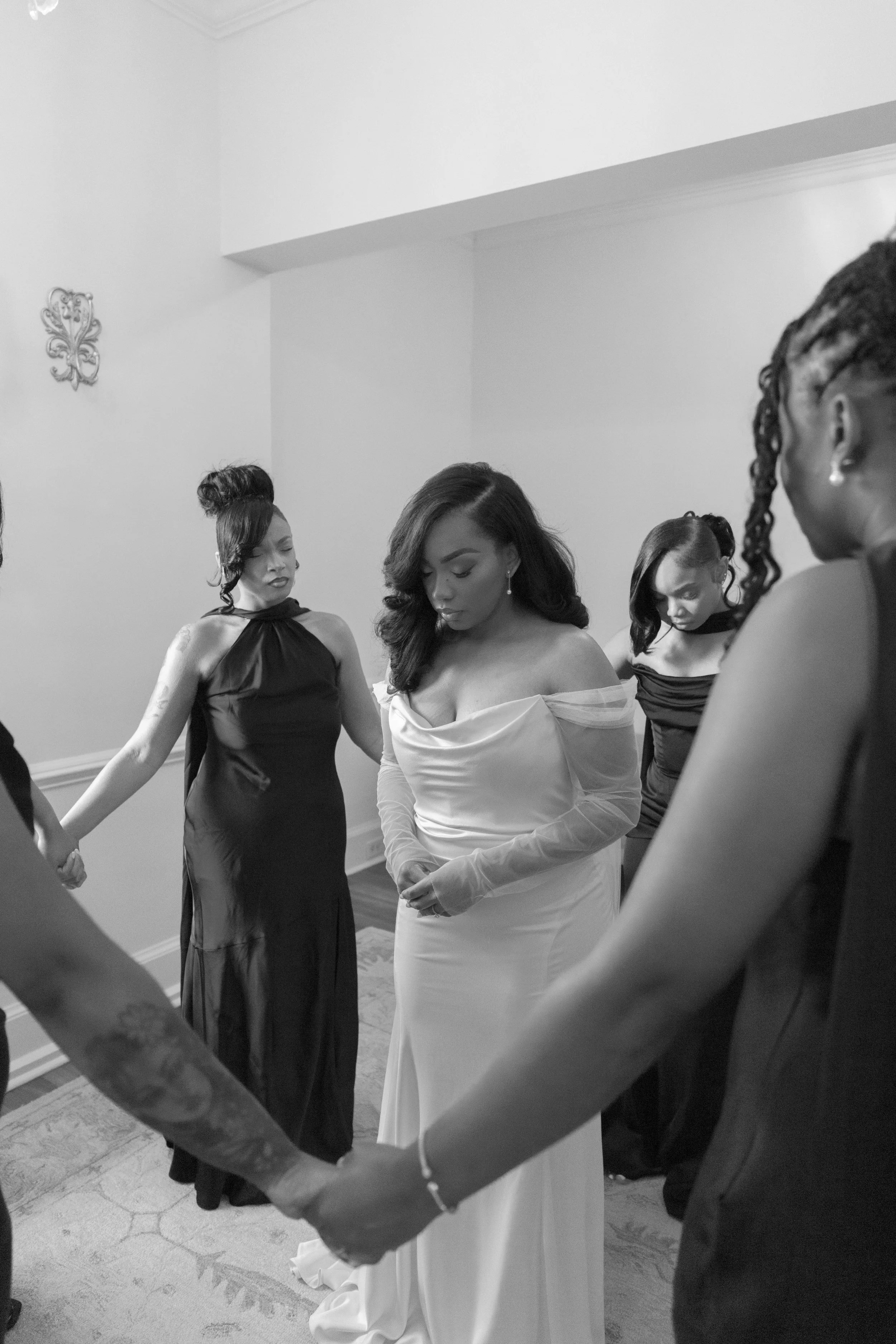 A group of women dressed in formal attire standing in a circle with their hands joined, engaged in a prayer or moment of reflection in a decorated room.