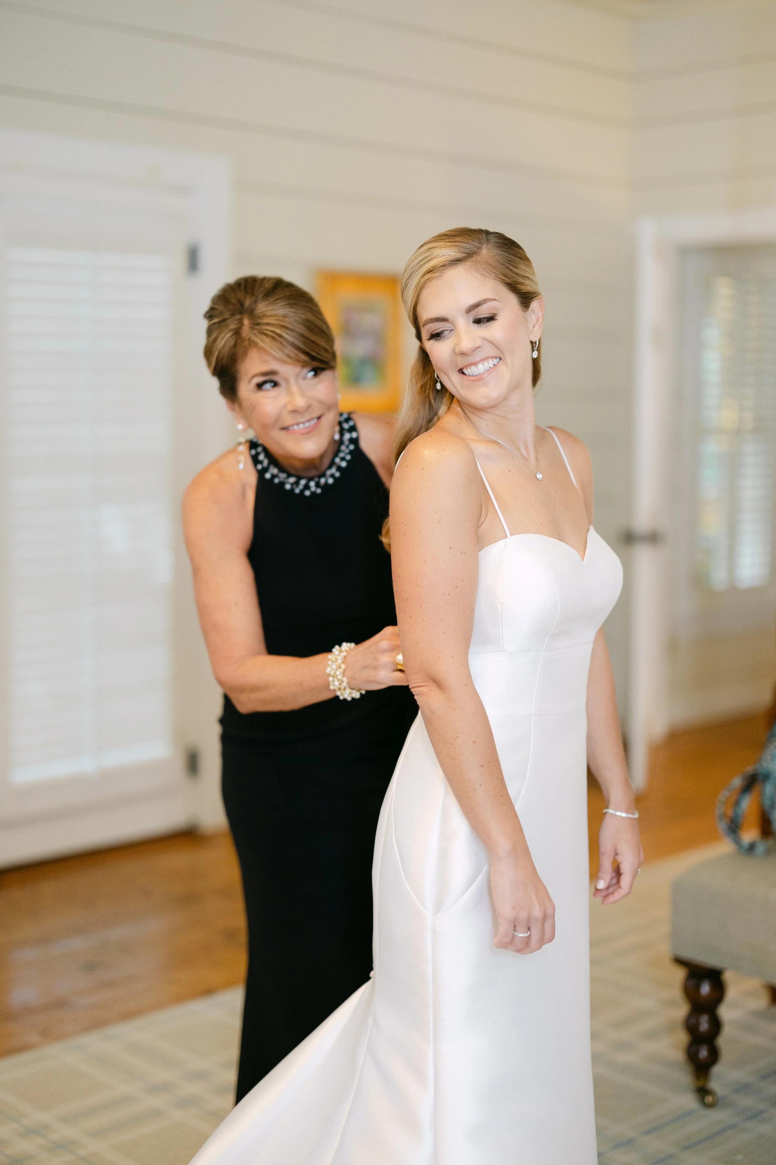 A bride in a white wedding dress smiling as someone helps her adjust the gown in a bright room.
