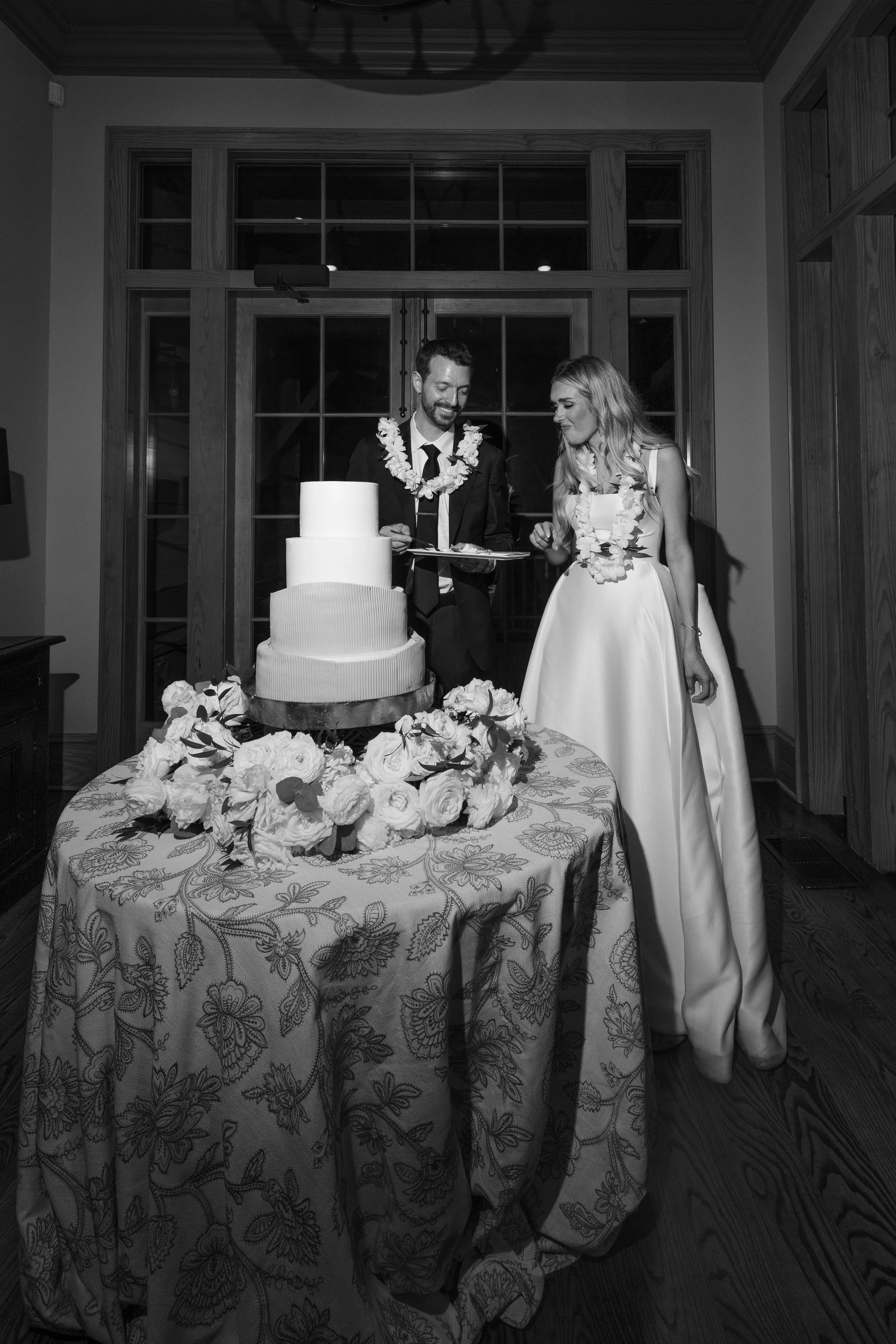 Black and white photo of a bride and groom with leis, standing next to a wedding cake on a decorated table, smiling at each other during their wedding celebration.