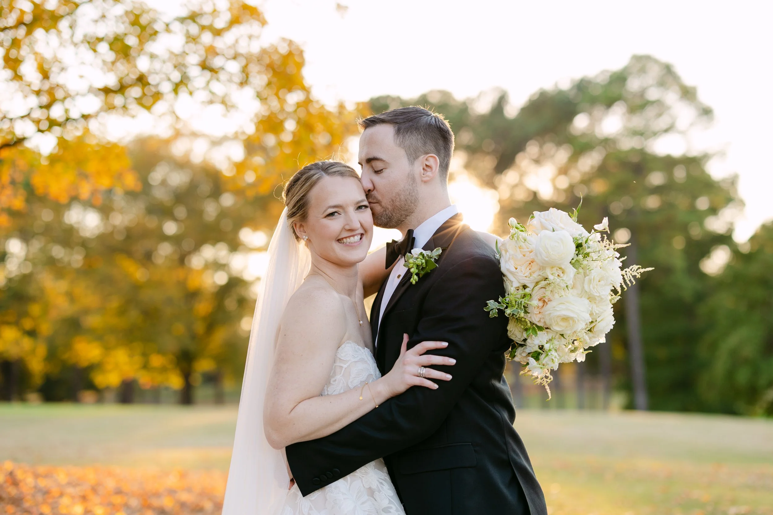 A newlywed couple sharing a kiss outdoors in a park during sunset, with the bride holding a bouquet of white flowers and both dressed in formal wedding attire.