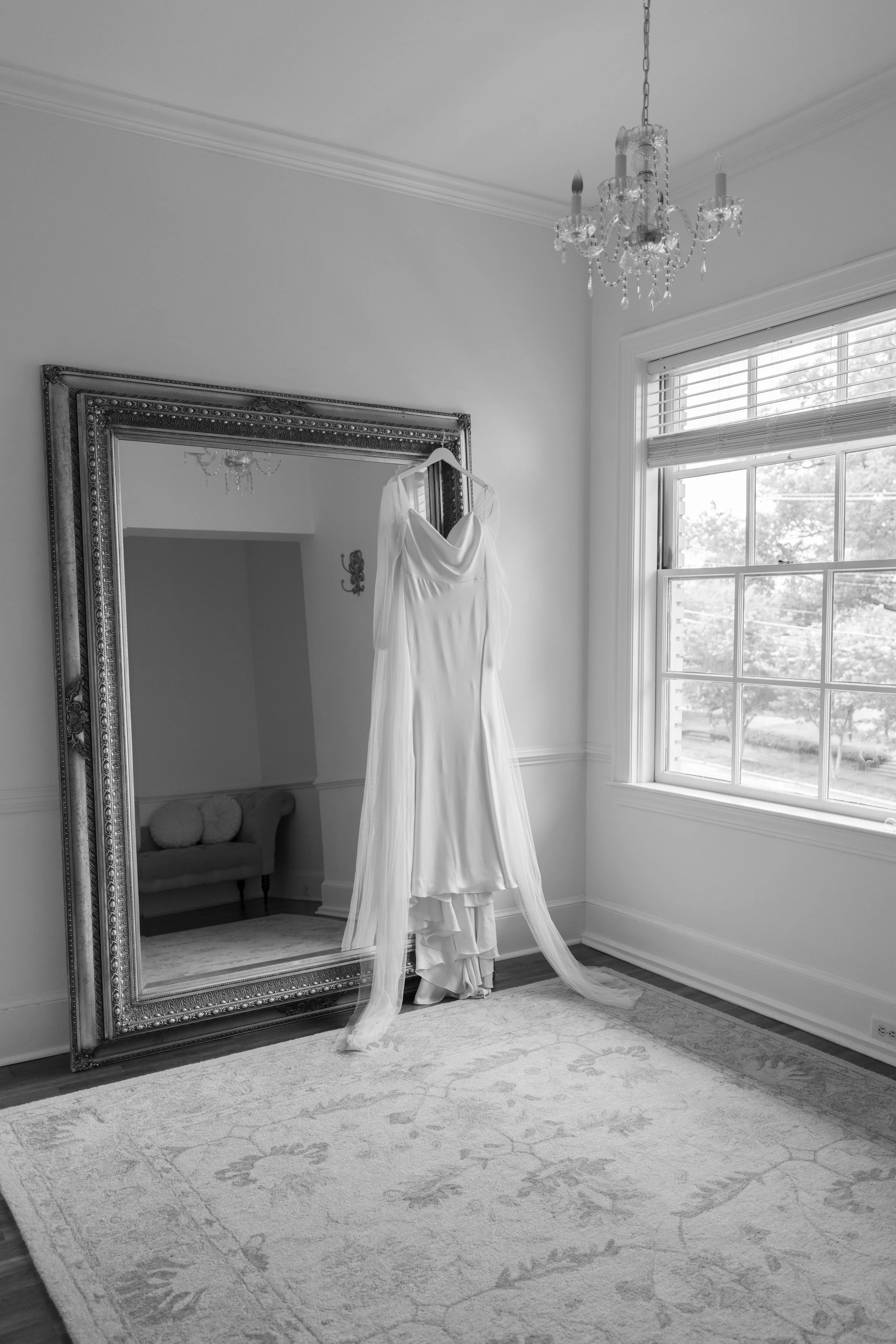 A white wedding dress hanging on a hanger in front of a large decorative mirror in a room with a window and a chandelier.
