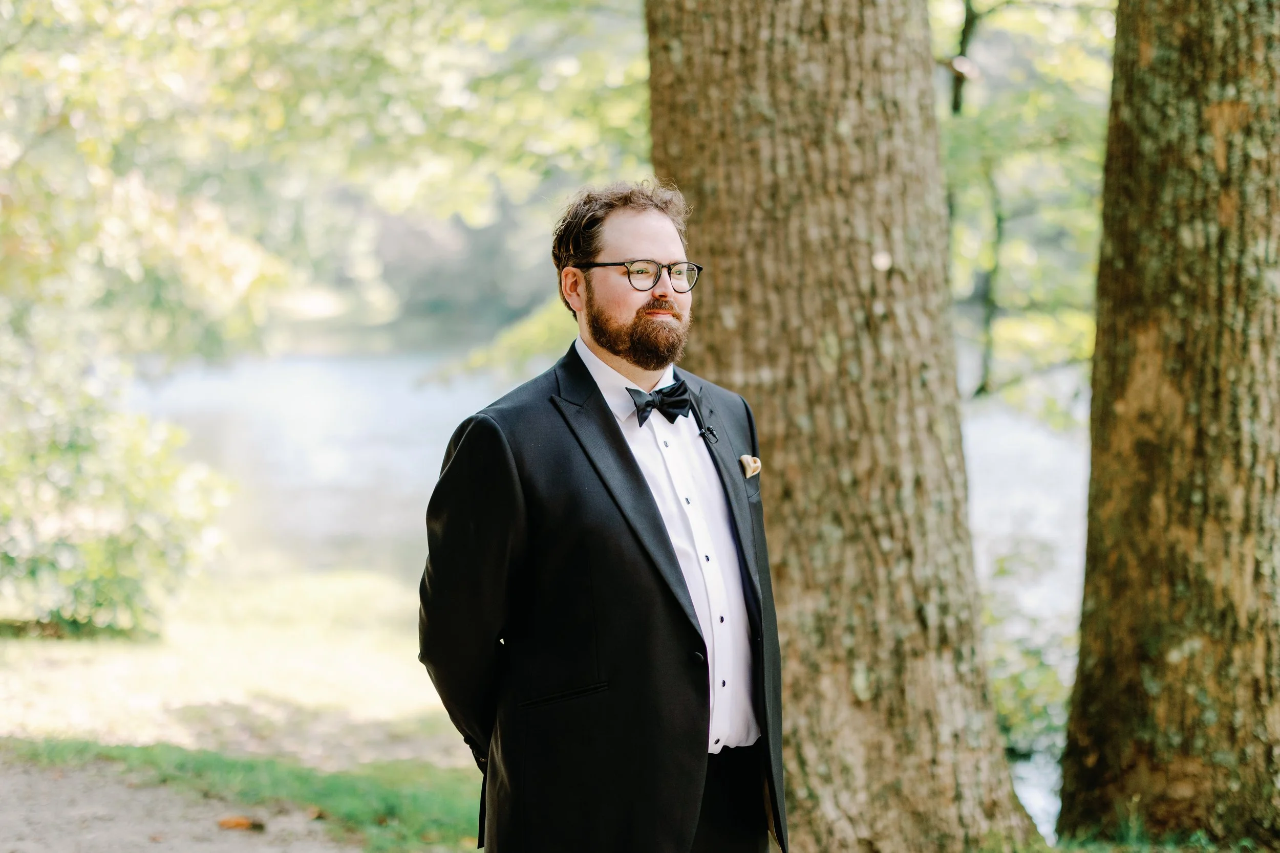A man dressed in a tuxedo with a bow tie standing outdoors near a large tree by a body of water, with trees and leaves in the background.