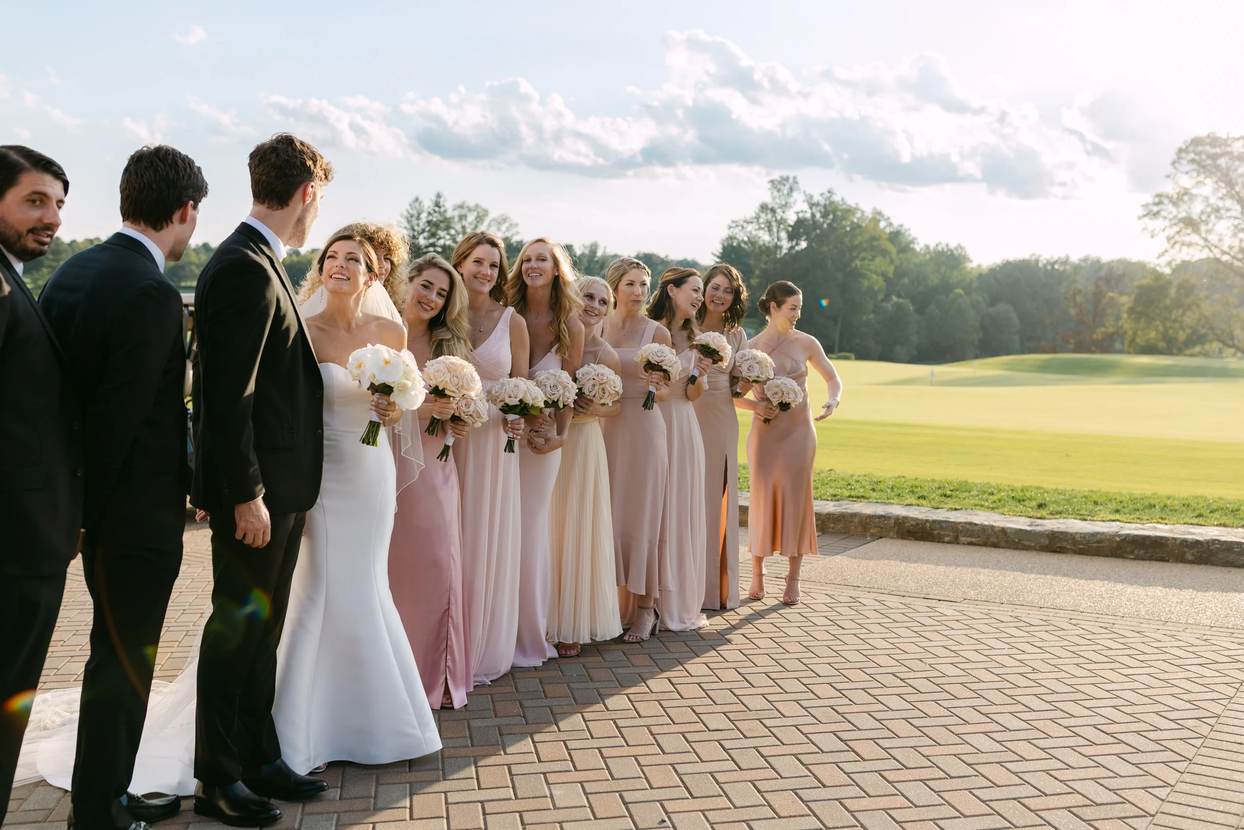 A bride and groom with a bridal party of women holding bouquets, standing outdoors on a brick path in a grassy area with trees and a cloudy sky in the background, during a wedding.