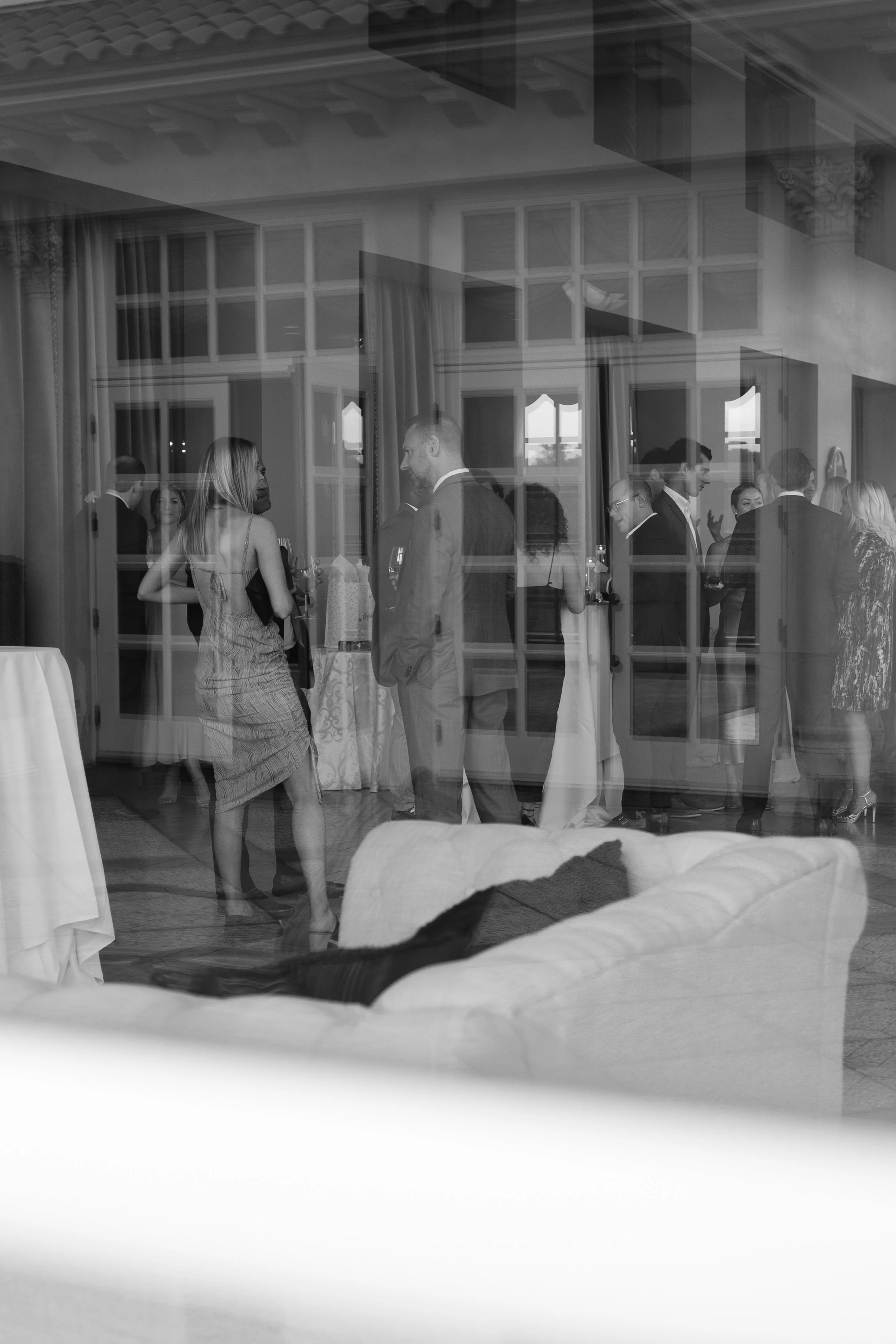 People socializing at an indoor event, seen through a glass window or door, in black and white.