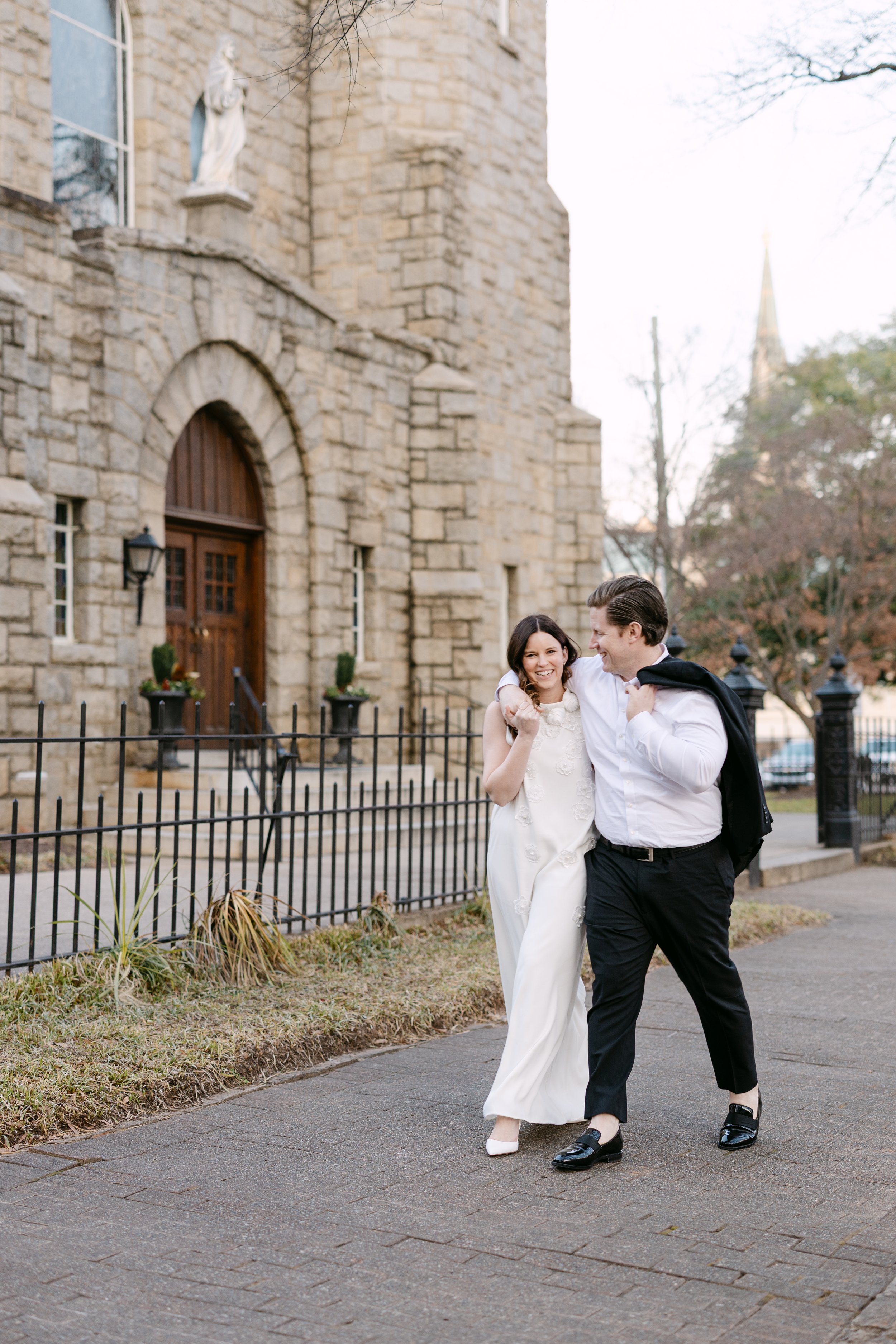 A couple walk arm in arm, smiling, outside a stone church, dressed in wedding attire, with autumn trees in the background.