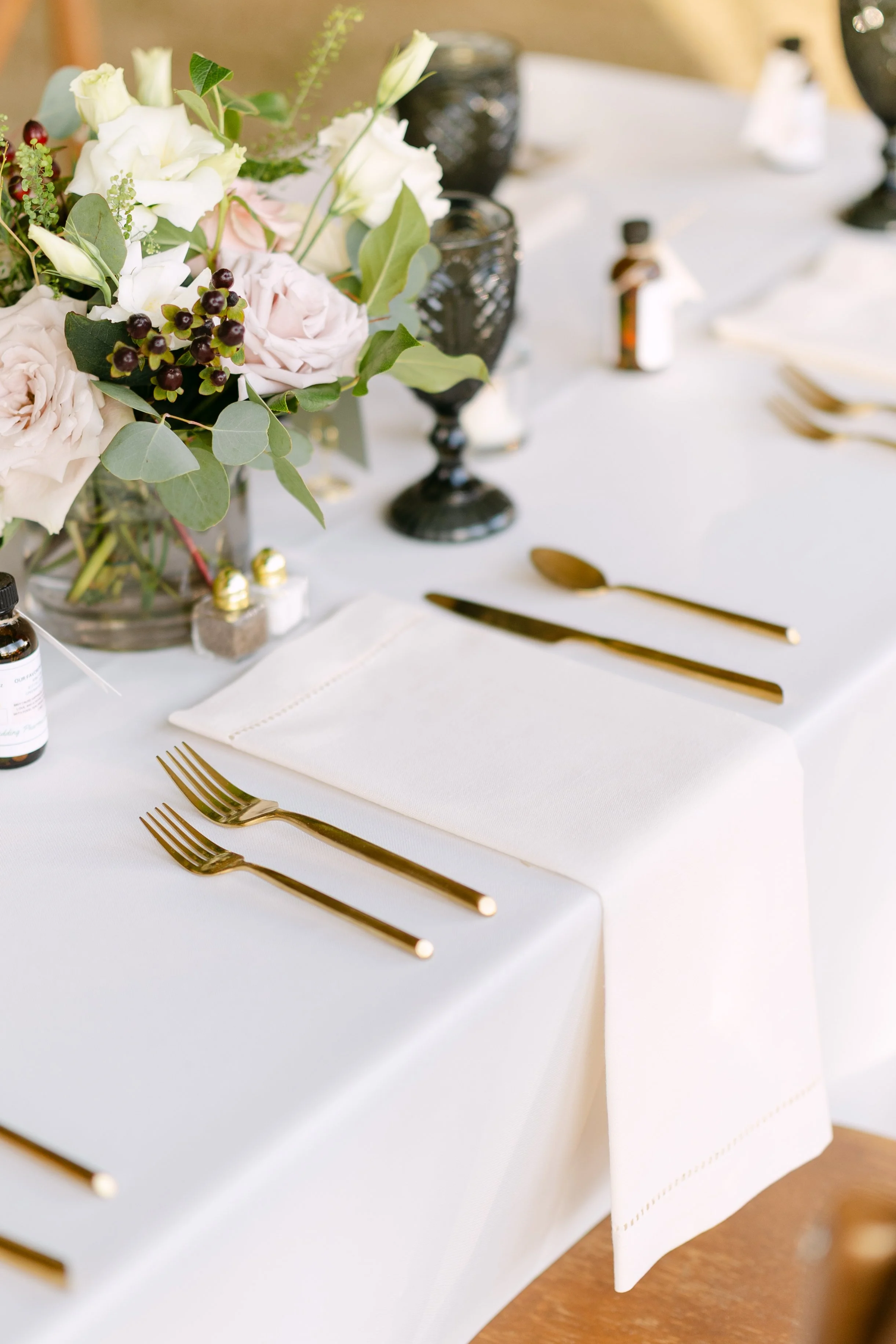 Elegant dining table setting with white napkins, gold cutlery, black goblets, and a floral centerpiece with white and blush roses, greenery, and dark berries.