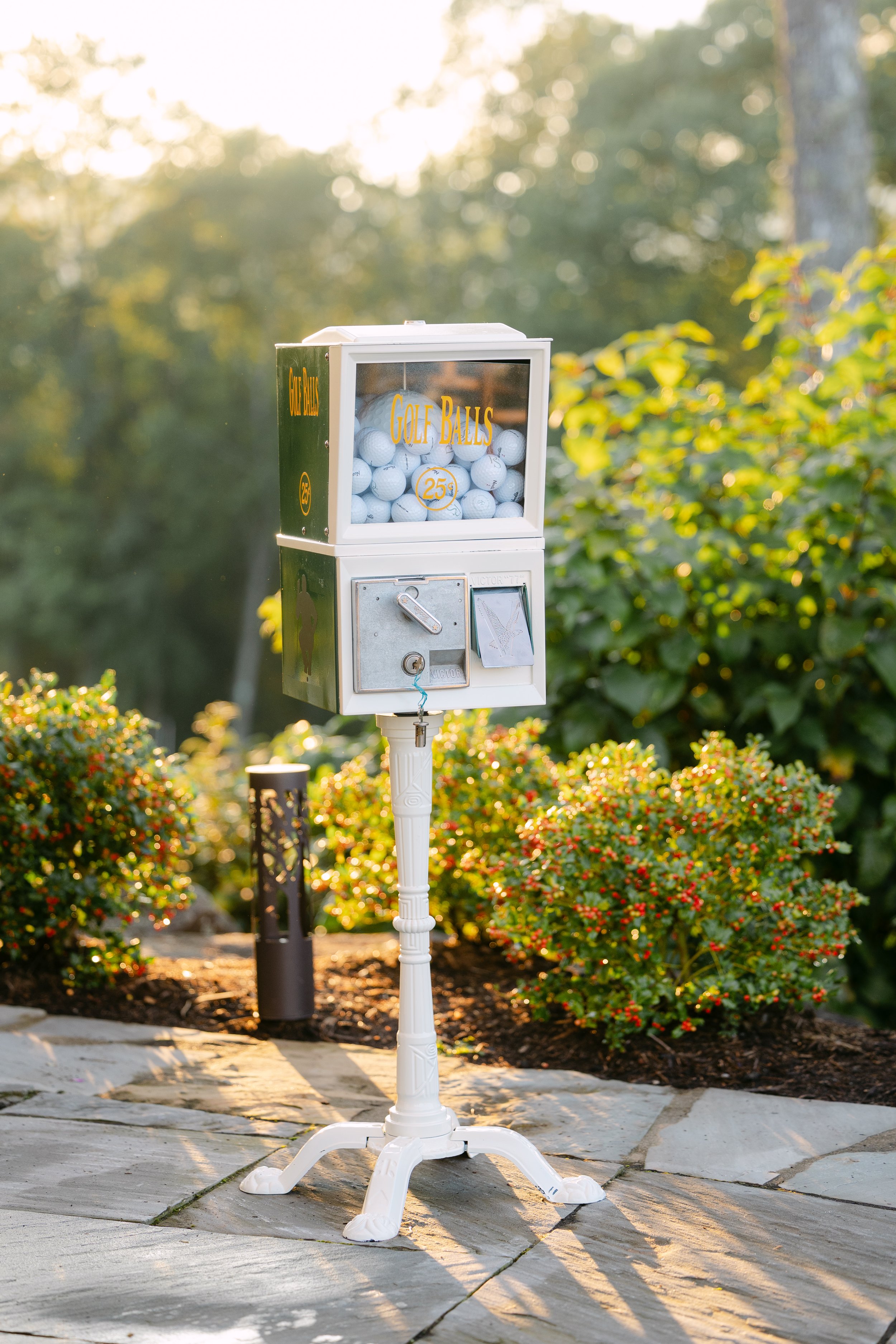 A vintage-style outdoor golf ball vending machine filled with white golf balls labeled "Golf Balls" and "25¢" stands on a white ornate pedestal. The machine is located on a stone patio, surrounded by green bushes and trees, with sunlight filtering th