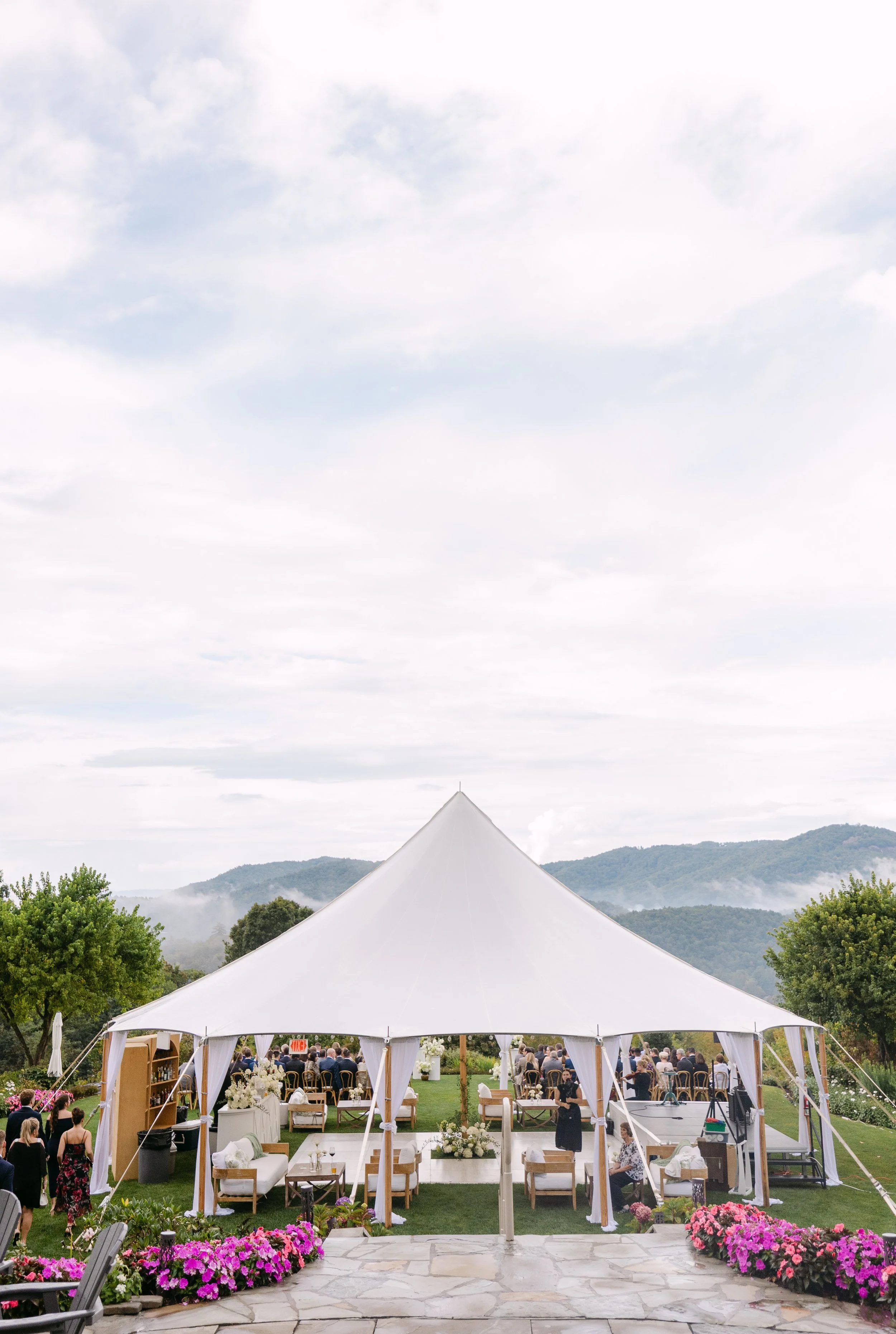 Outdoor wedding reception under a large white tent with guests seated at tables, surrounded by colorful flowers and scenic mountains in the background.