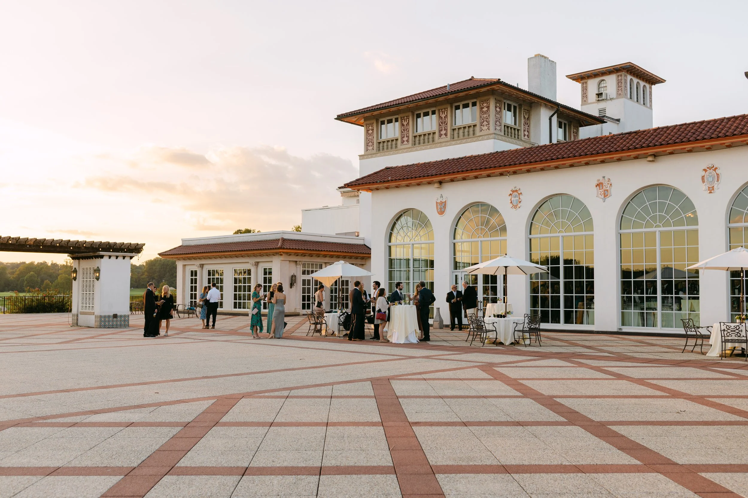 People gathered at an outdoor event on a terrace of a large white building with arched windows, umbrellas, and tables