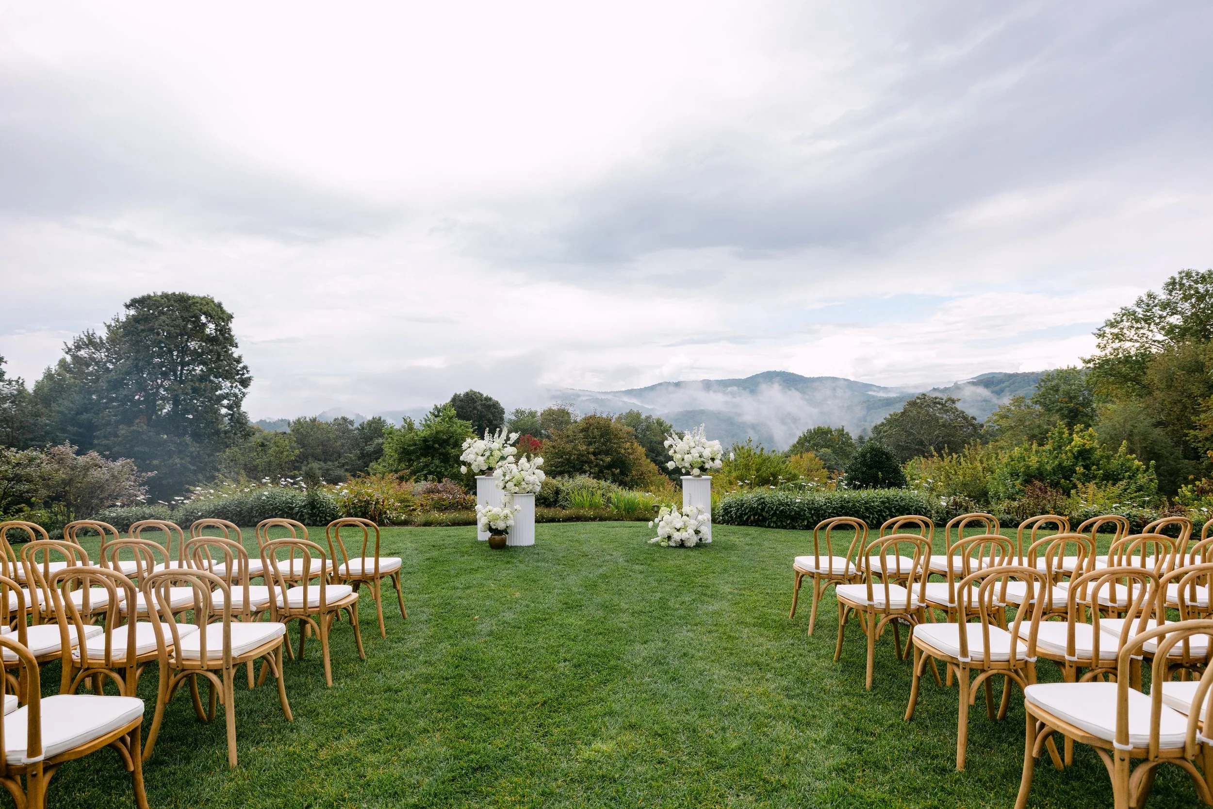 Outdoor wedding ceremony setup on a grassy lawn with rows of wooden chairs with white cushions, decorated with large white floral arrangements on pedestals, against a backdrop of trees, mountains, and cloudy sky.