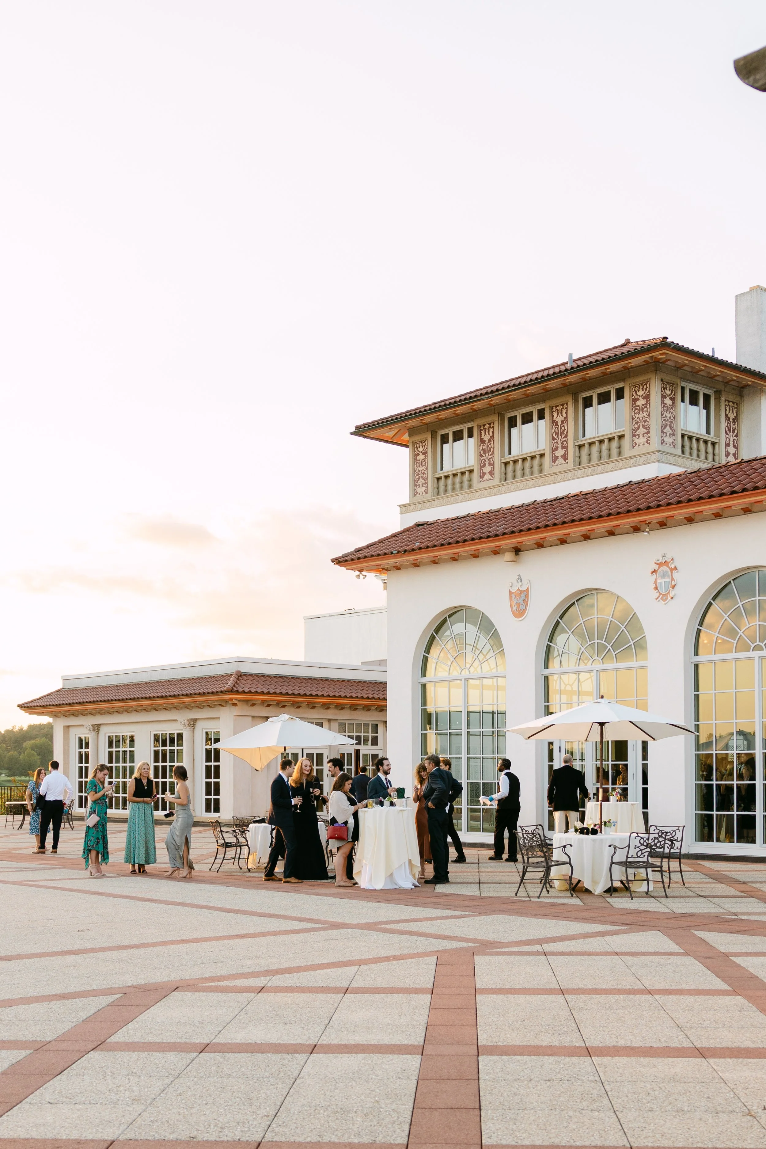 Guests attending a formal outdoor event on the terrace of a large white building with tall arched windows and decorative shields. Some guests are standing around tables with white tablecloths and umbrellas, mingling and holding drinks, with a sunset 