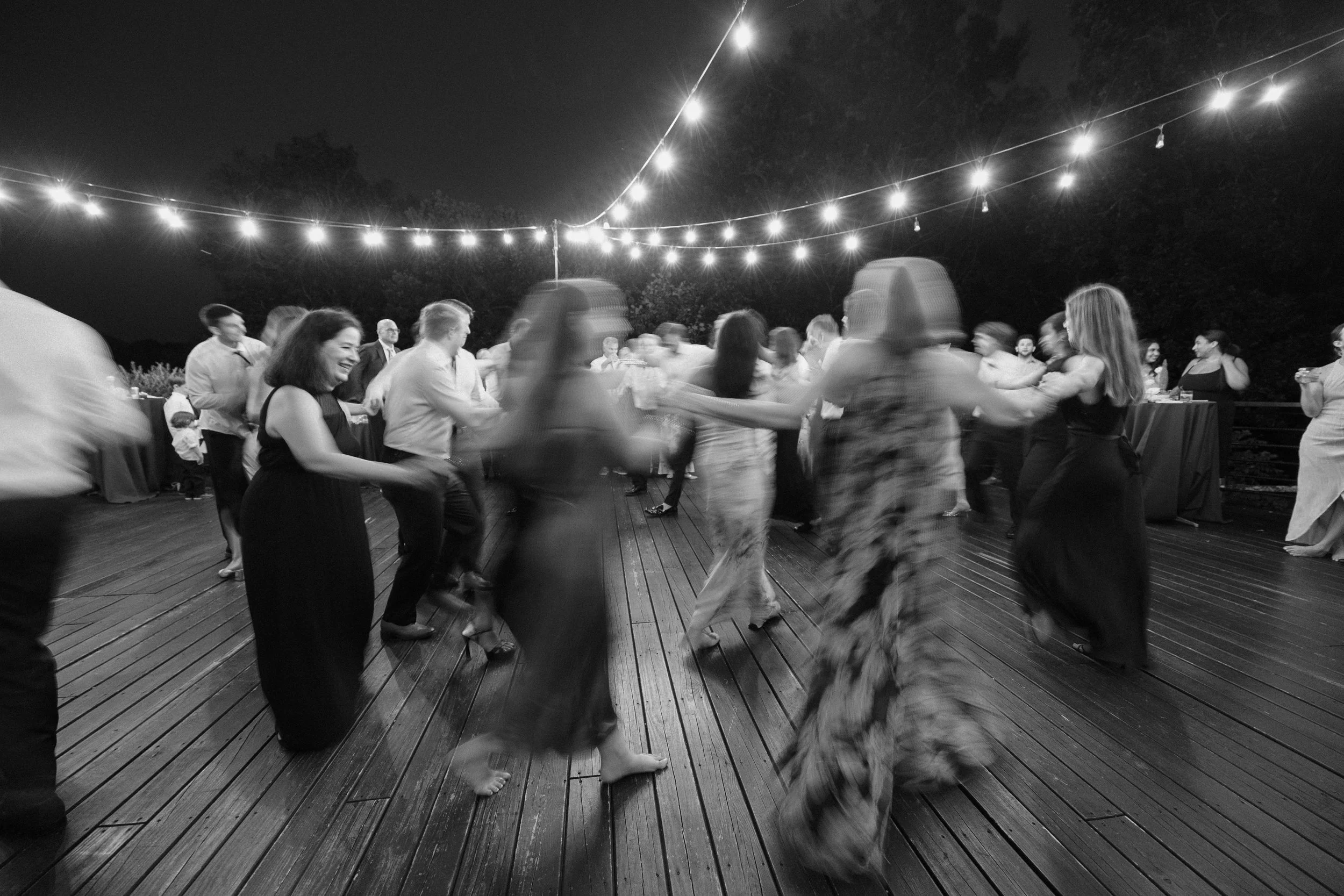 People dancing at an outdoor party under string lights at night, with the photo in black and white and some motion blur.
