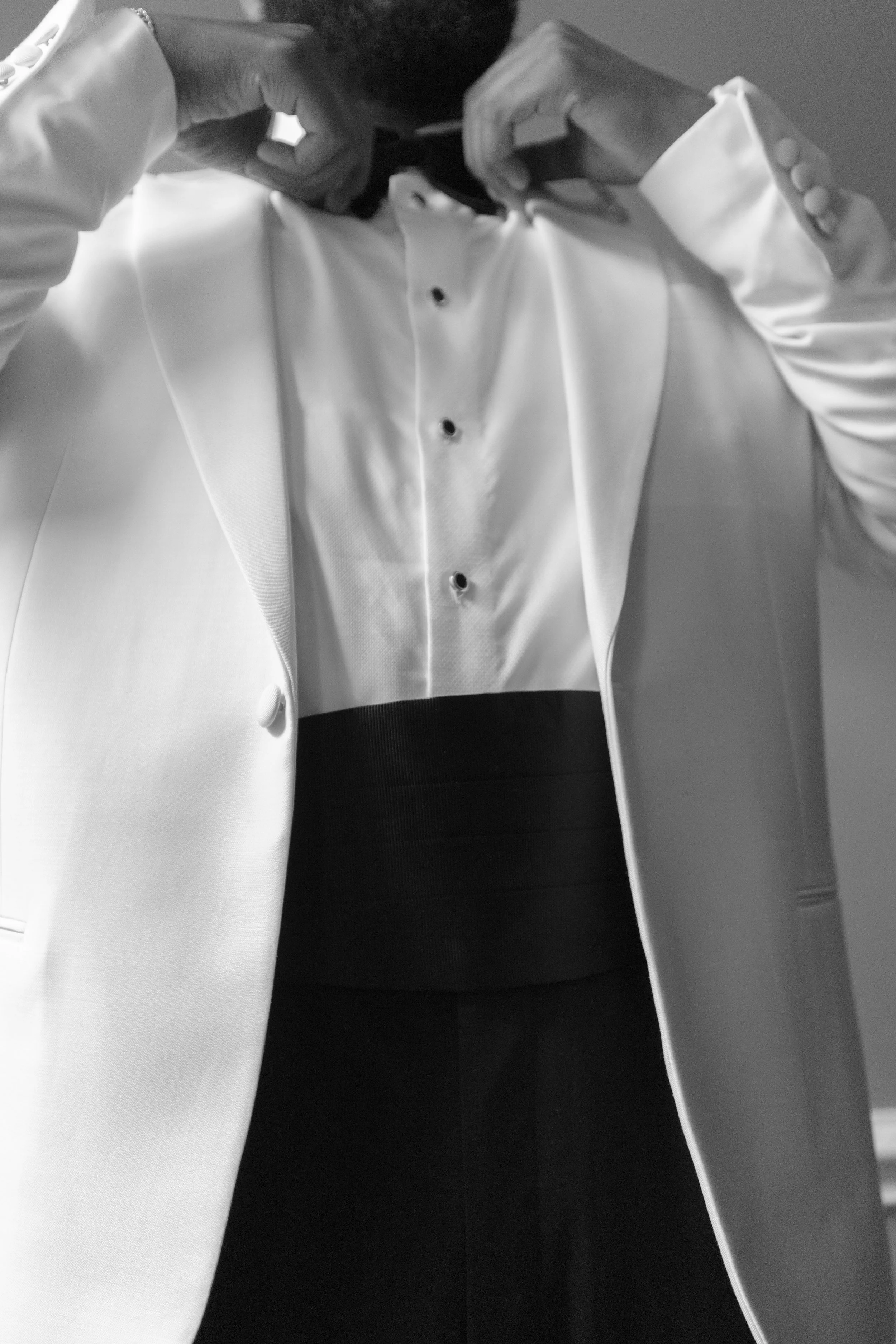 A person in a tuxedo with a white jacket and black shirt, adjusting their bowtie.