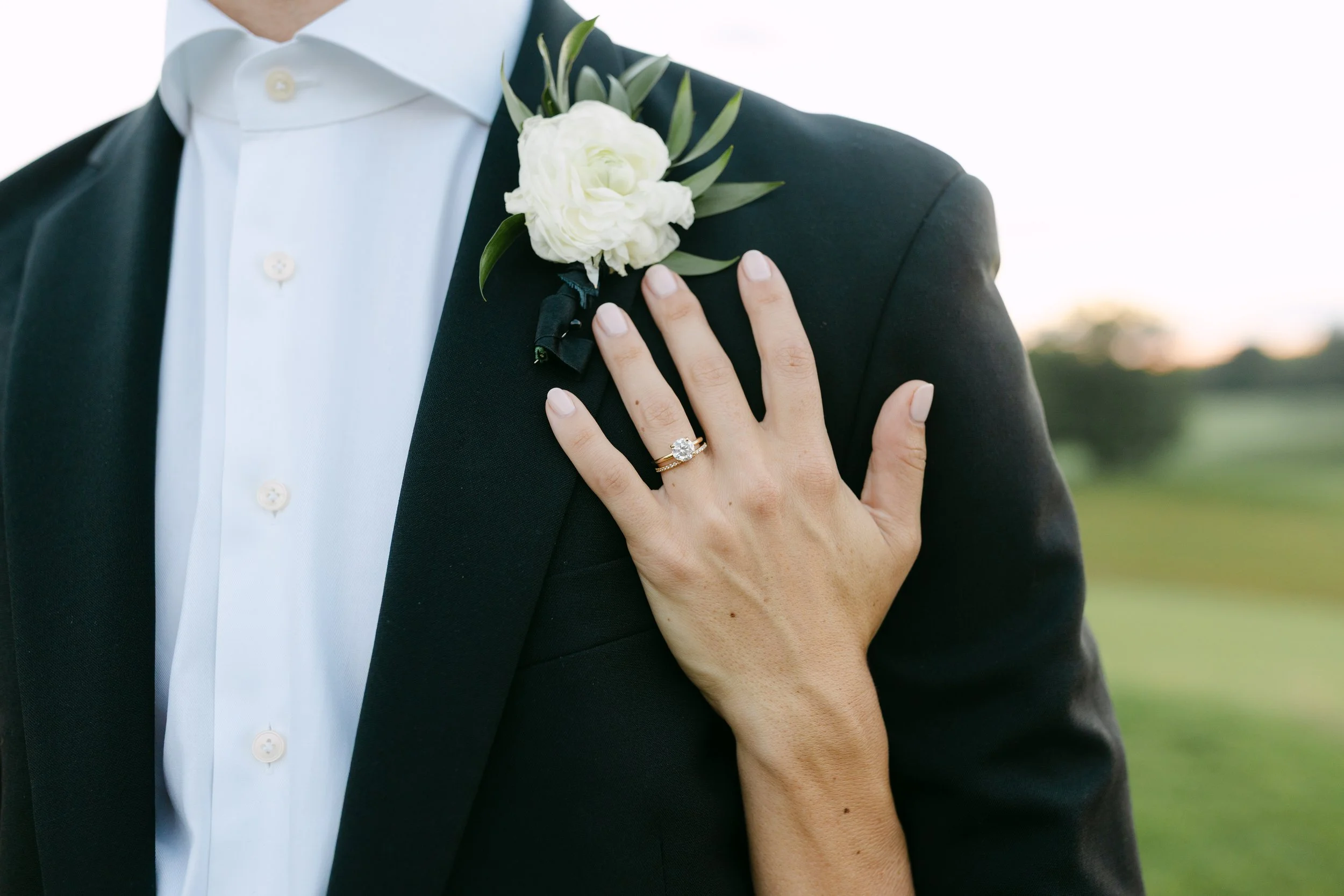 Person wearing a black tuxedo with a white shirt and a boutonniere on the left lapel, showing a hand with a ring, standing outdoors