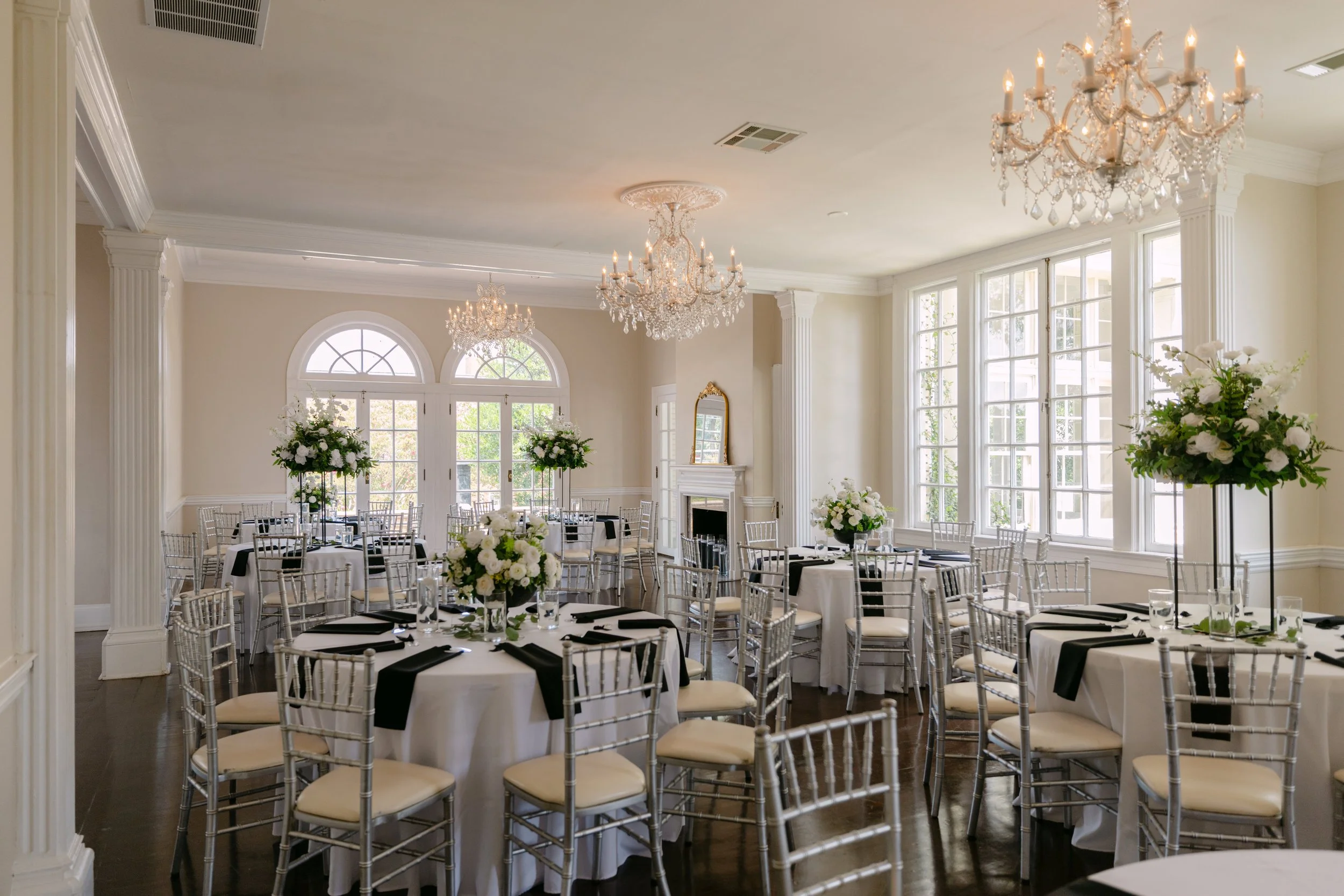 Elegant banquet hall decorated for a wedding reception with round tables covered in white tablecloths, black napkins, floral centerpieces with white flowers, and silver chairs. The room features large windows, a white fireplace, and ornate chandelier