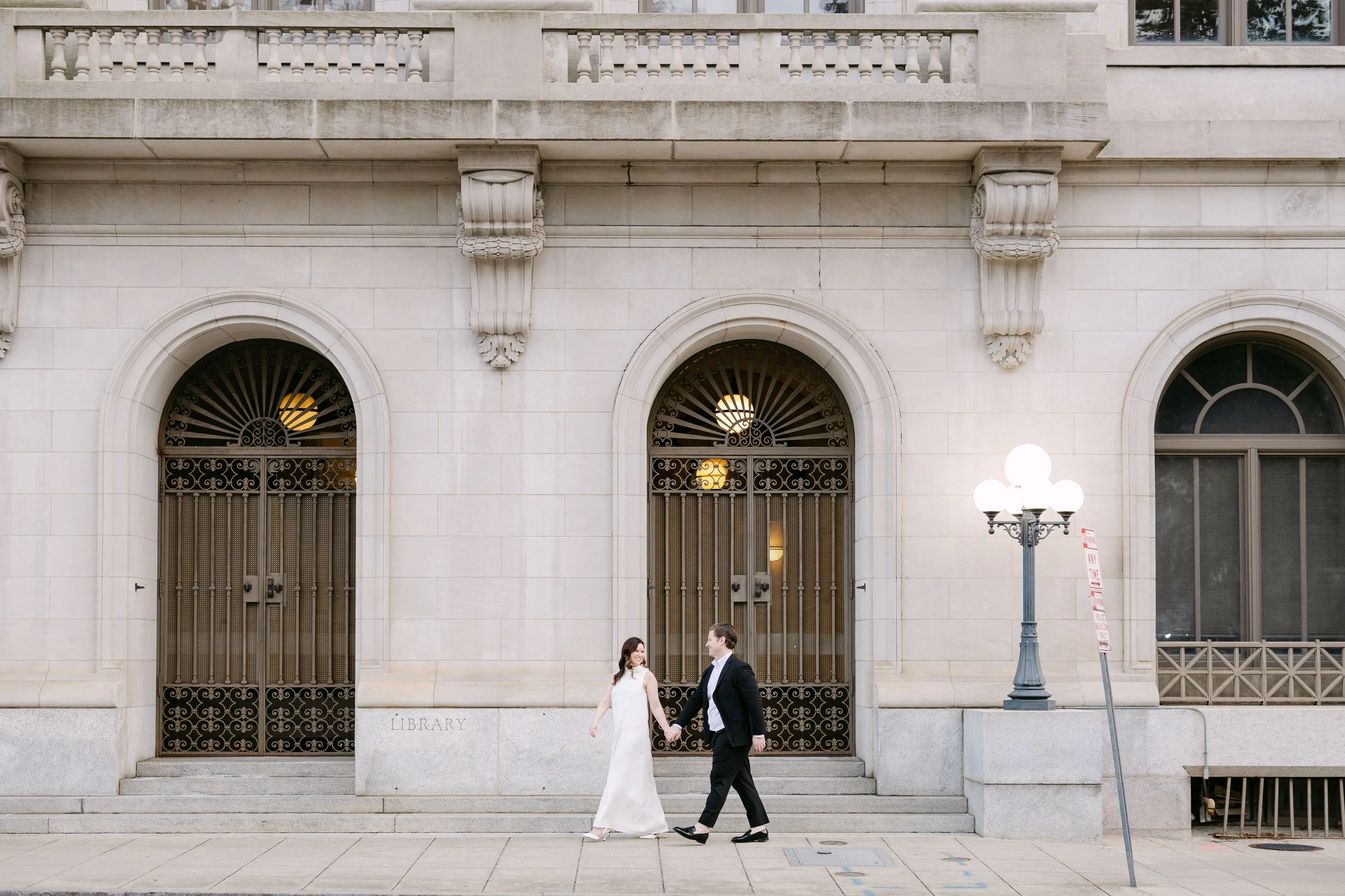 A couple walking hand in hand outside a stone library building with large arched windows and ornate iron gates.