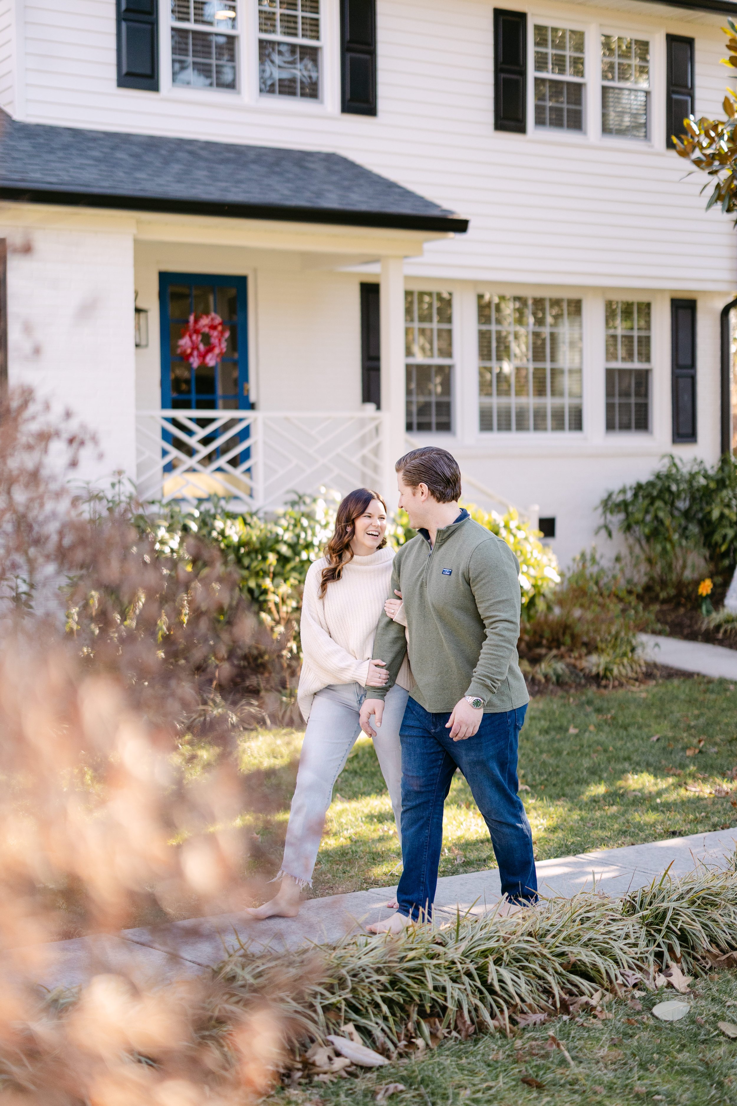 A couple walking barefoot on a sidewalk in front of a house, smiling and looking at each other, with greenery and flowers around them in a residential neighborhood.