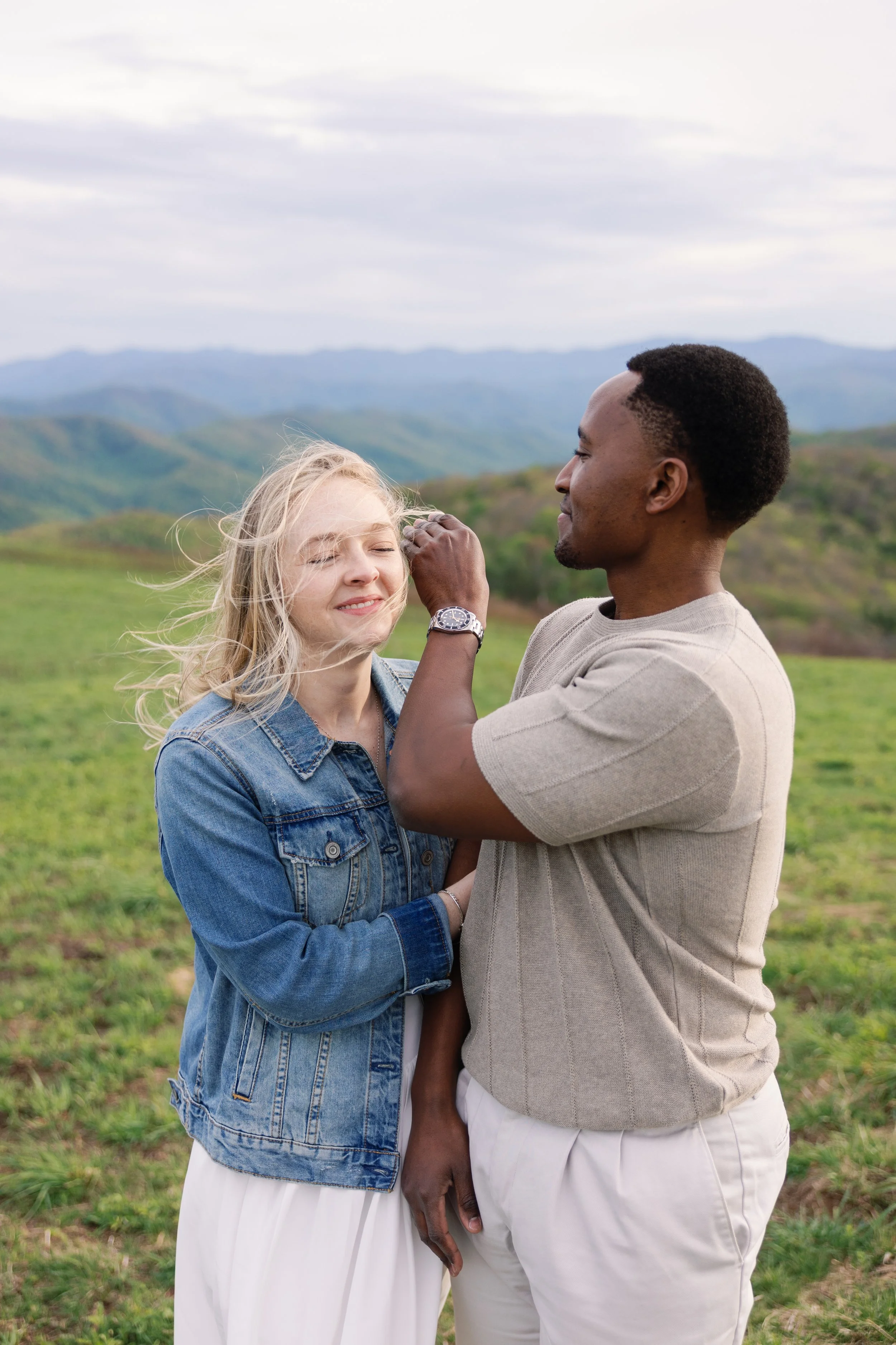 A man and a woman standing outdoors in a grassy field with hills and mountains in the background. The man is touching the woman's face, and both are smiling with eyes closed, sharing a tender moment.