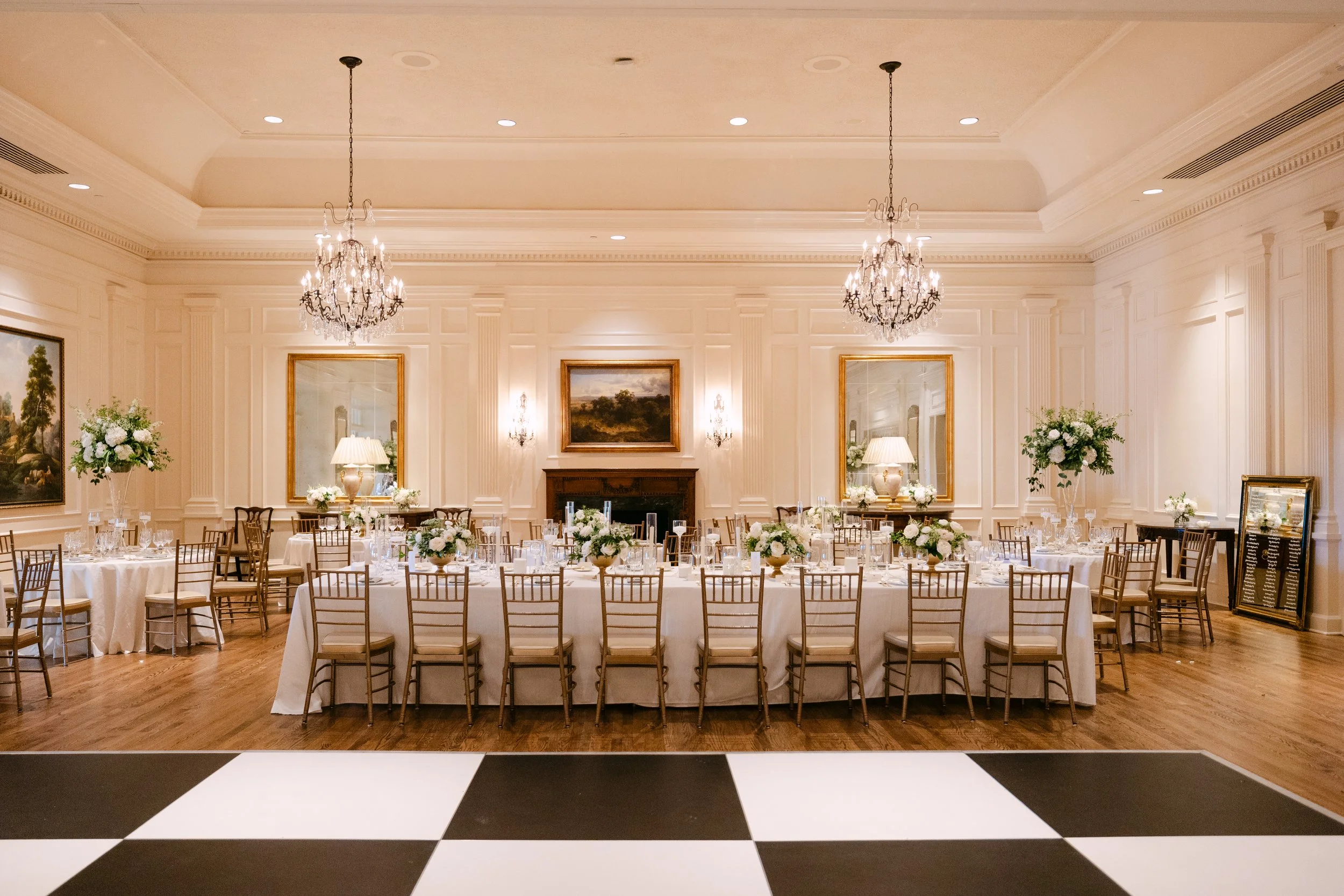 Elegant banquet hall with white walls, crystal chandeliers, and framed artwork. Long dining table with white tablecloth, floral centerpieces, and place settings, surrounded by wooden chairs. Wooden floor, black and white checkered dance floor in fore