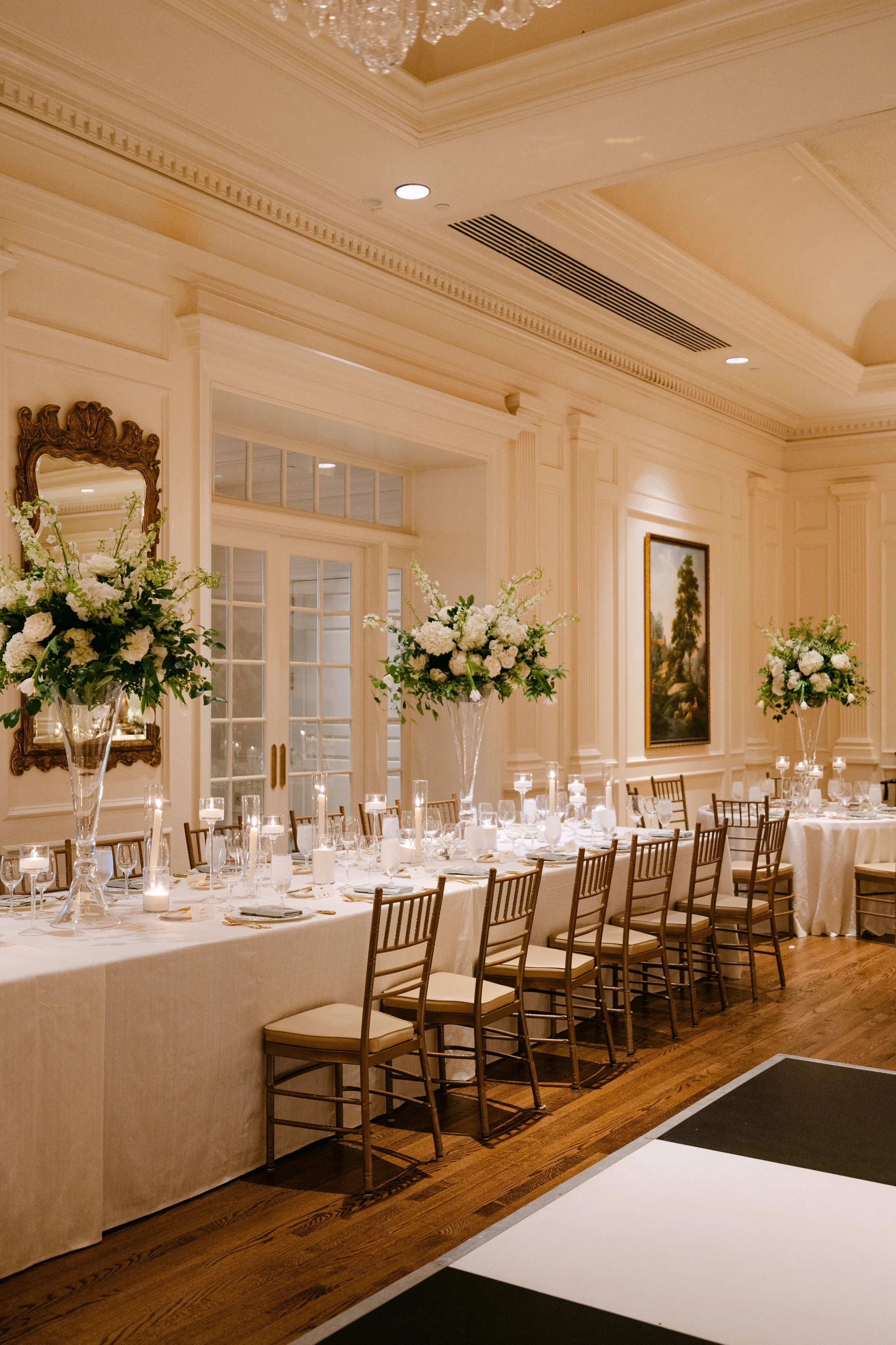 Elegant dining room with a long table, floral centerpieces, candles, and chairs, set for a formal event in a cream-colored room with artwork and large windows.