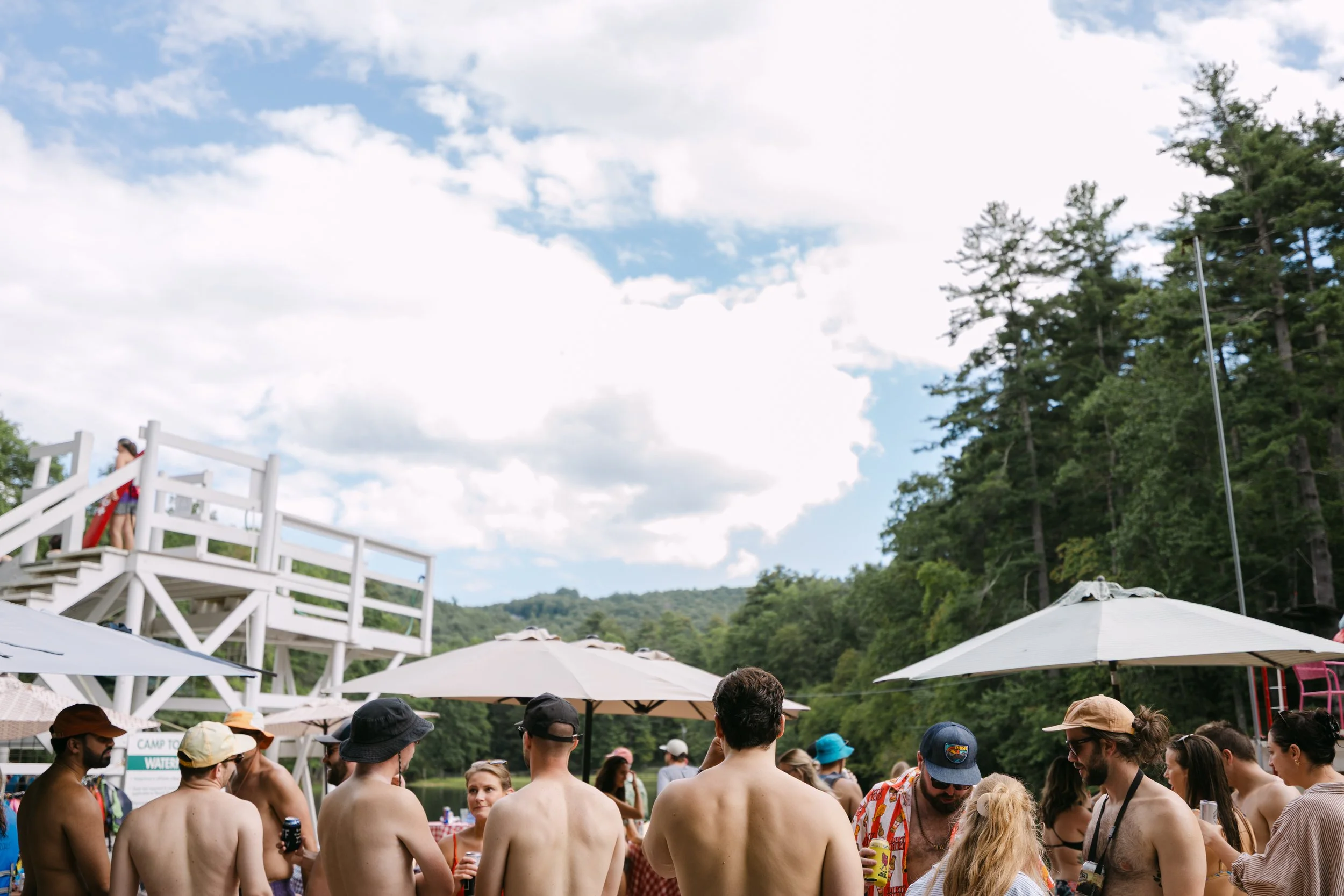 A group of people in swimsuits and summer clothing gathering outdoors near a lake, with an observation deck on the left and trees in the background, under a partly cloudy sky.