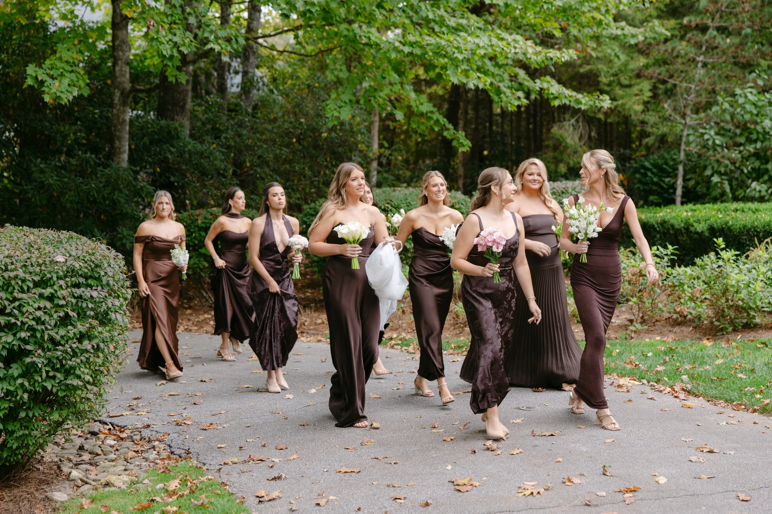 Bride and bridesmaids in dark brown dresses carrying bouquets while walking through a wooded garden.