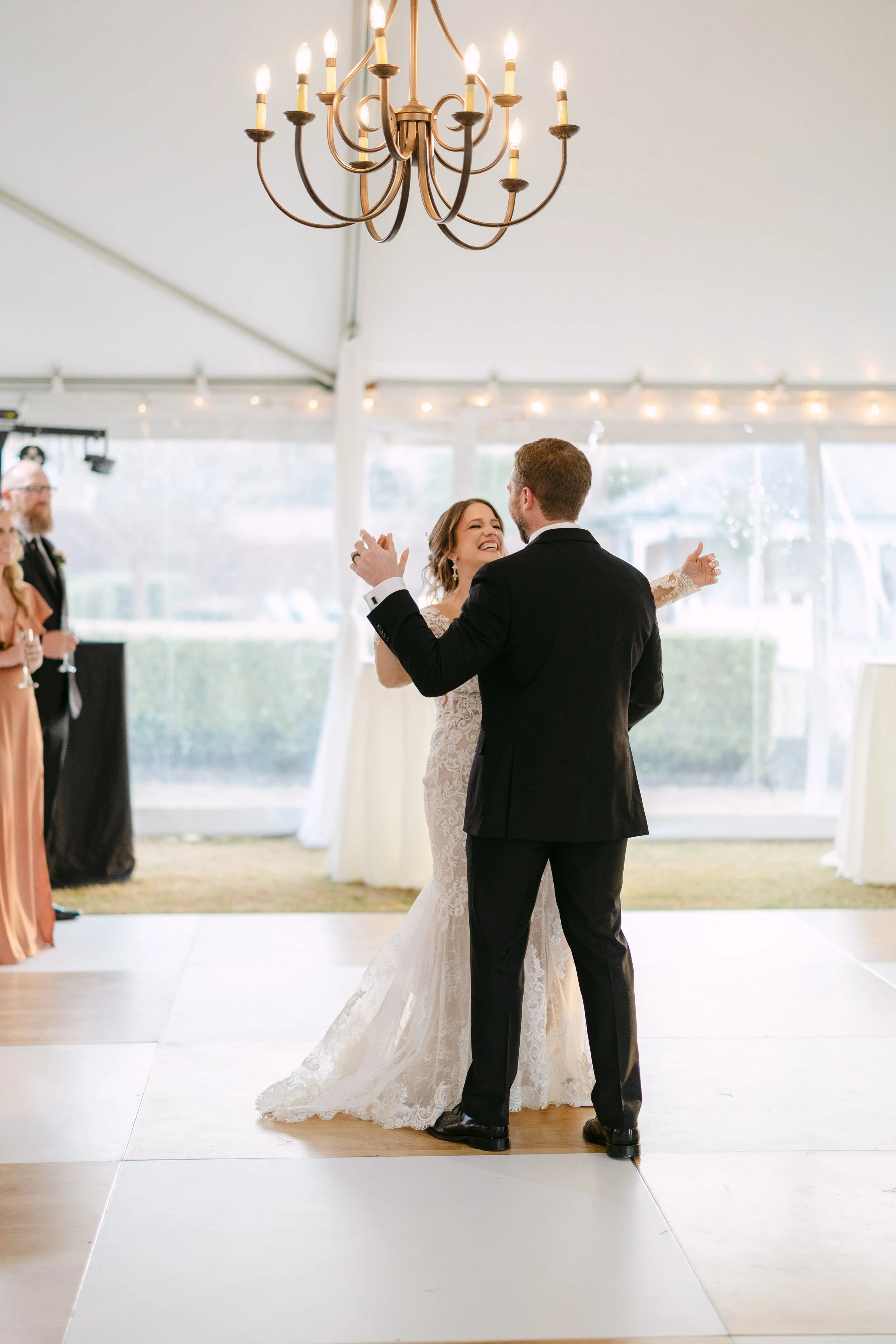 A bride and groom dancing at their wedding reception inside a tent, with guests watching in the background. The bride is wearing a white lace wedding dress and the groom is in a black suit.