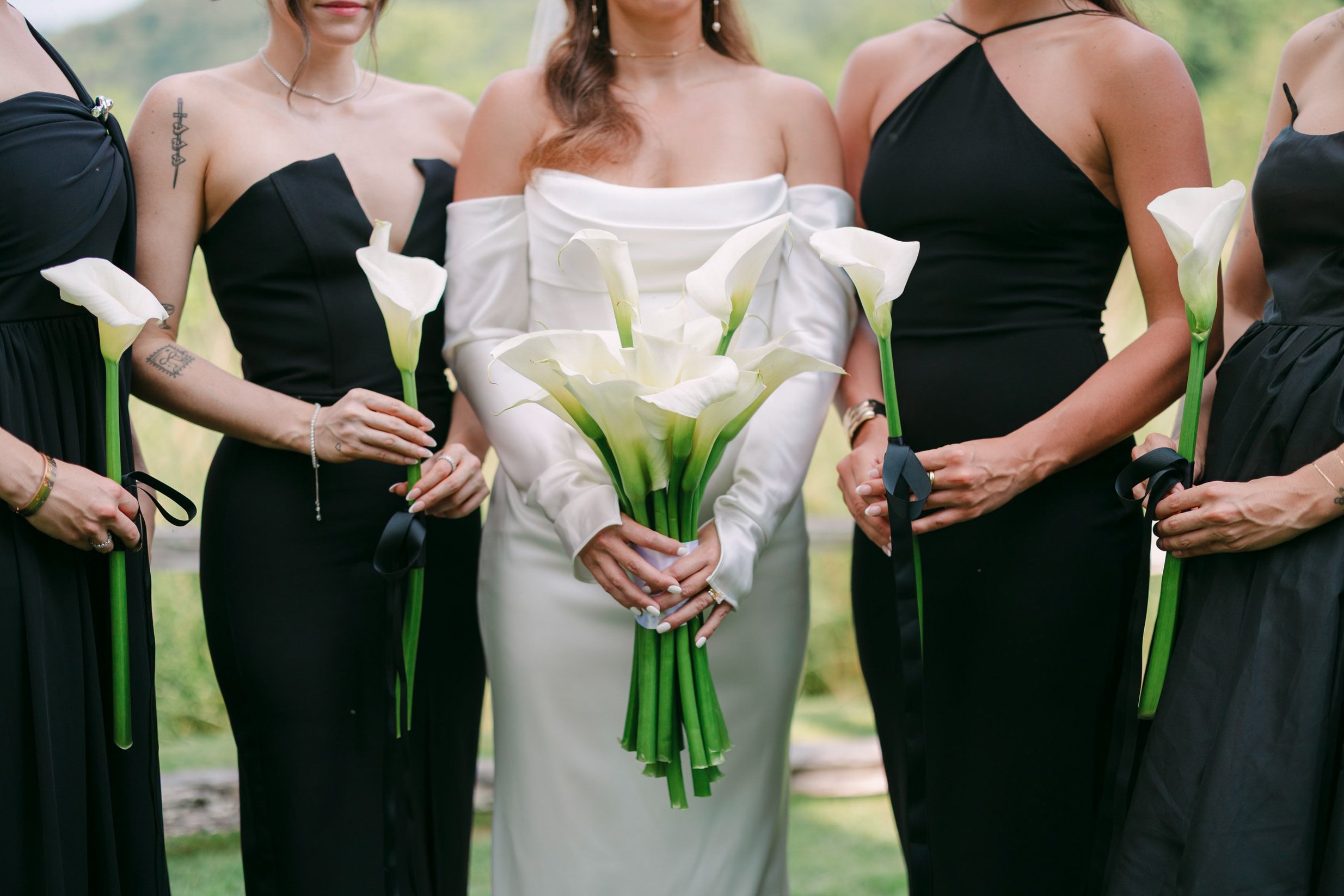 Group of women at a wedding, with the bride in a white off-shoulder dress holding a bouquet of calla lilies, and bridesmaids in black dresses holding similar calla lilies.