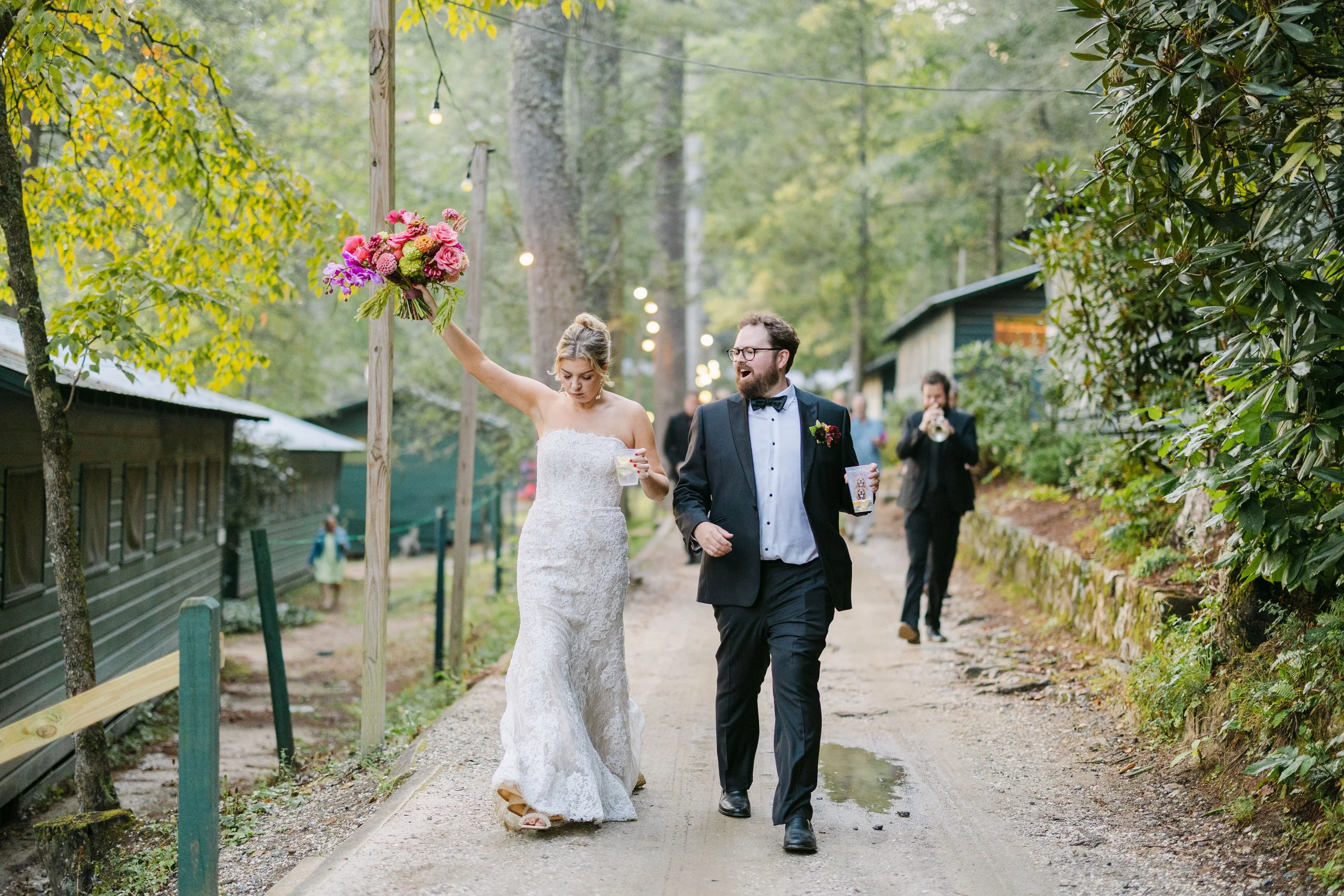 Bride and groom walking on a dirt path in outdoors, with the bride holding a bouquet raised in the air and the groom smiling, while a musician plays trumpet in the background.