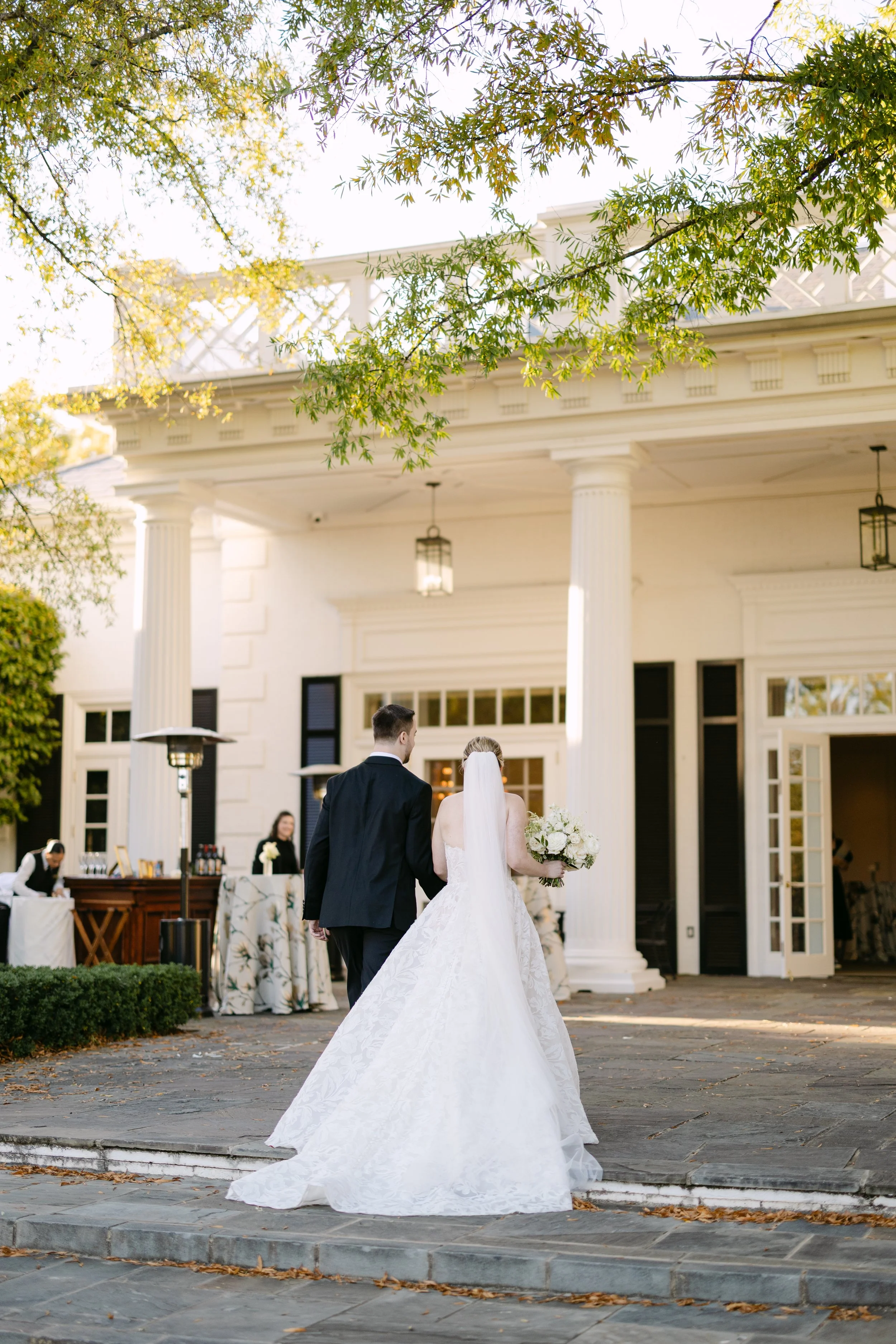 Bride and groom walking away from the camera outside a white building with large columns, during a wedding ceremony with greenery overhead.