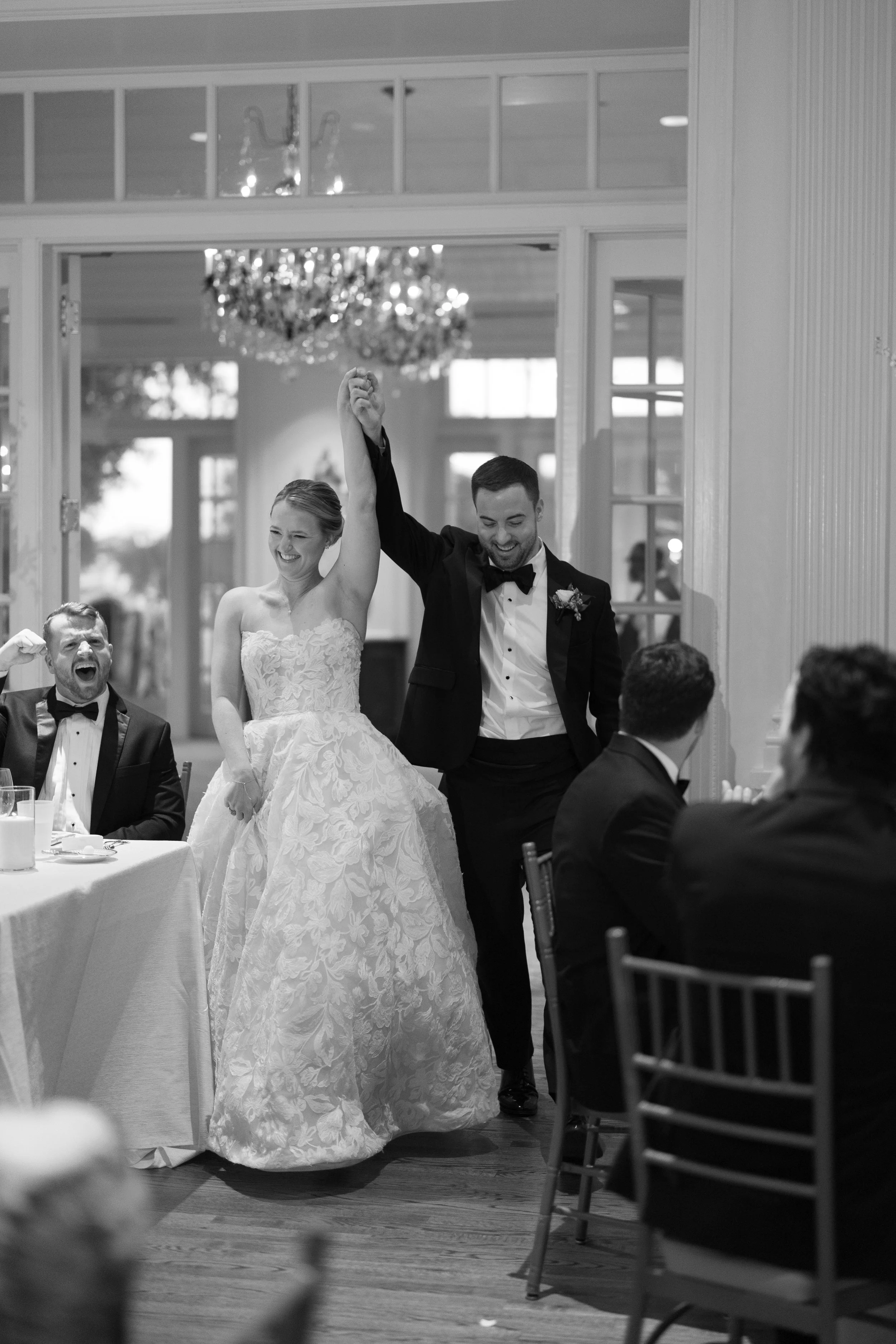 A newlywed couple, the bride and groom, celebrating with friends and family at a wedding reception. The groom is lifting the bride's arm in a victory pose, and everyone is smiling and laughing.