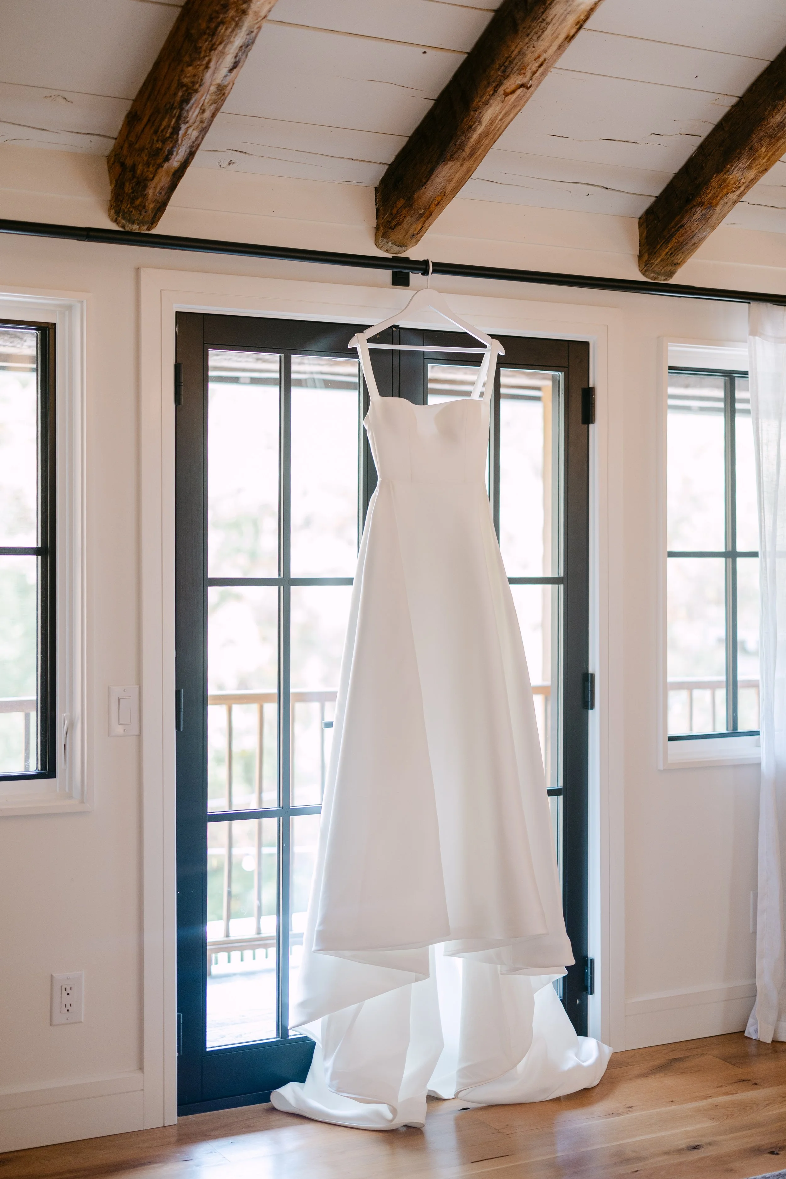 White wedding dress hanging on a hanger in front of a glass door with windows, with wooden beams on the ceiling and sunlight coming through the windows.