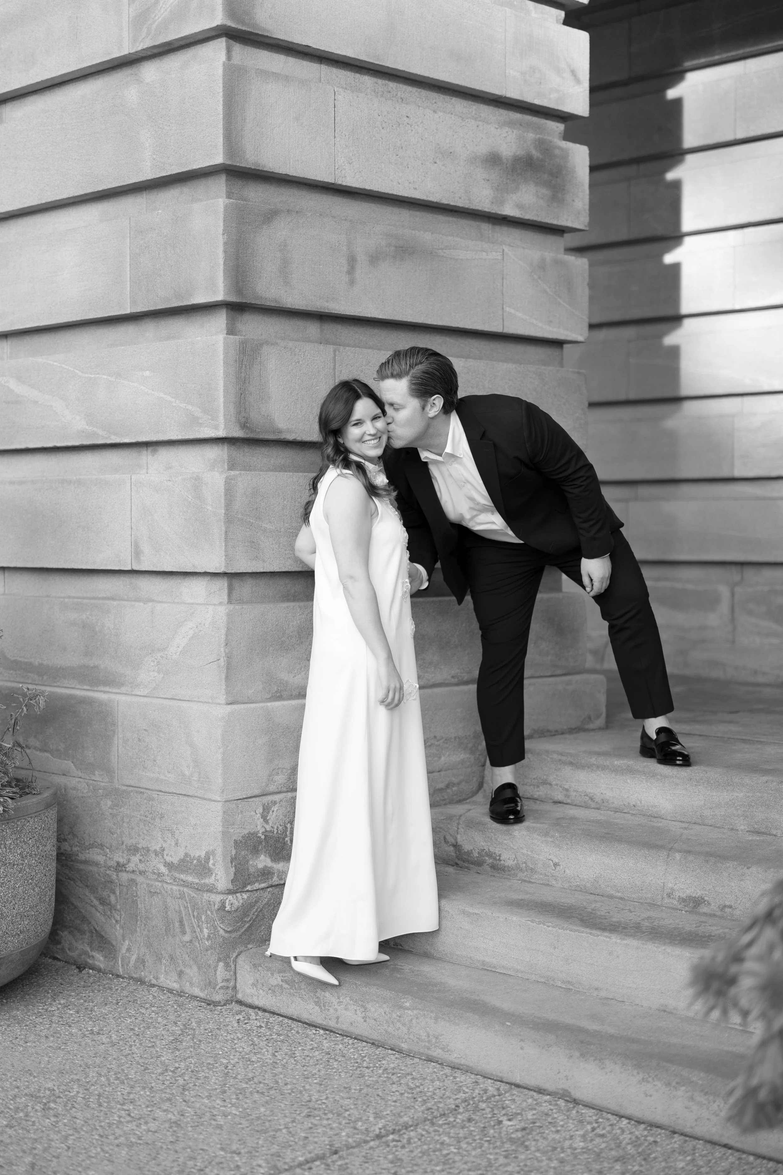 A man in a suit kisses a woman in a white dress on the cheek outside a building.