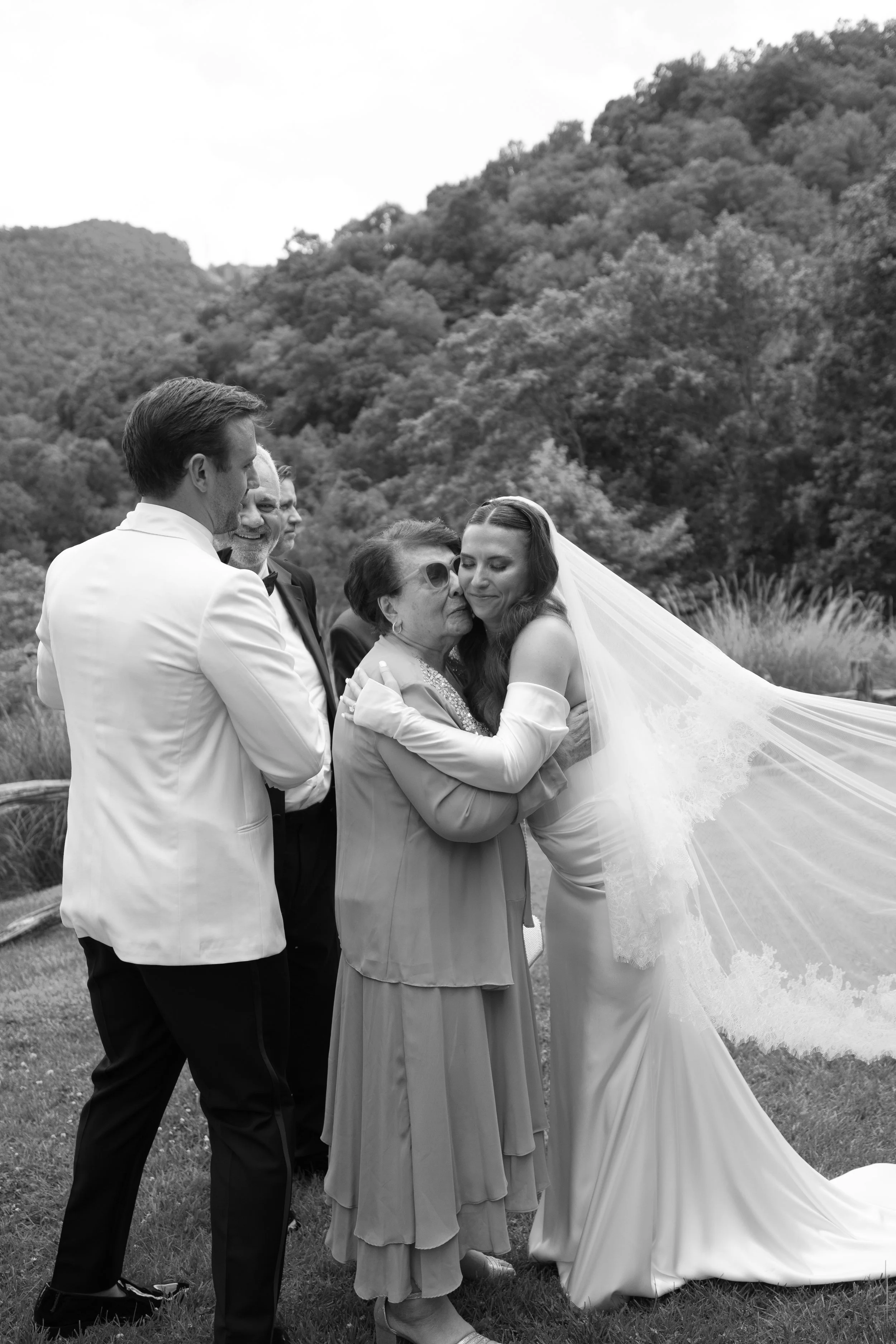 A bride in a wedding gown and veil hugging an older woman with sunglasses, surrounded by four men in tuxedos outdoors with a wooded hill in the background.
