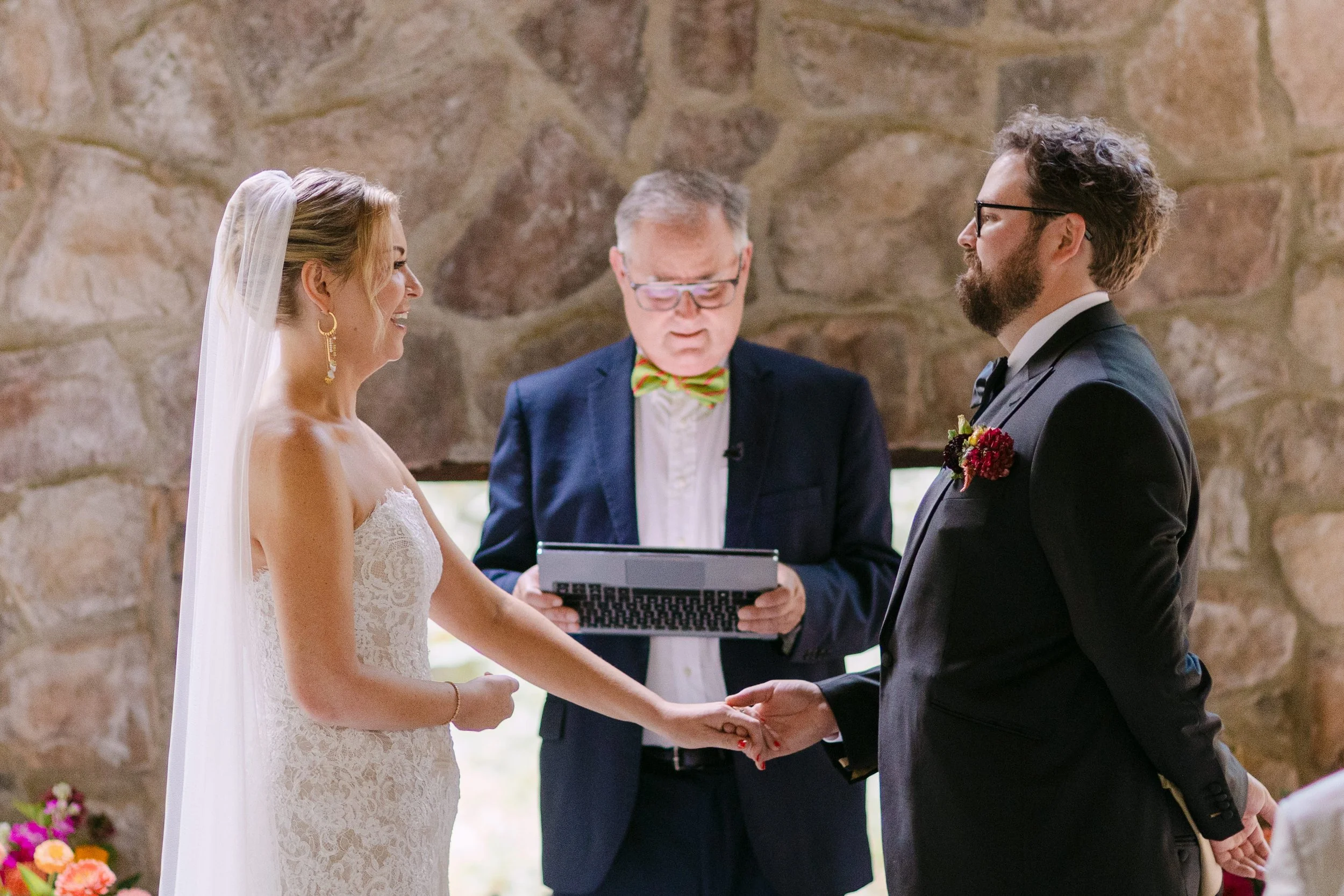 A bride and groom exchange wedding vows while holding hands, with an officiant reading from a tablet in a rustic indoor setting.