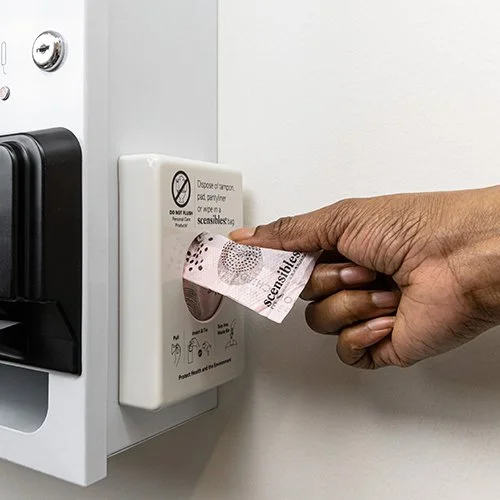 Person taking a menstrual care disposal bag from a compact dispenser attached to the side of a tampon and sanitary pad free vending machine in order to discard in a safe, sanitary and discreet way in a women's restroom.
