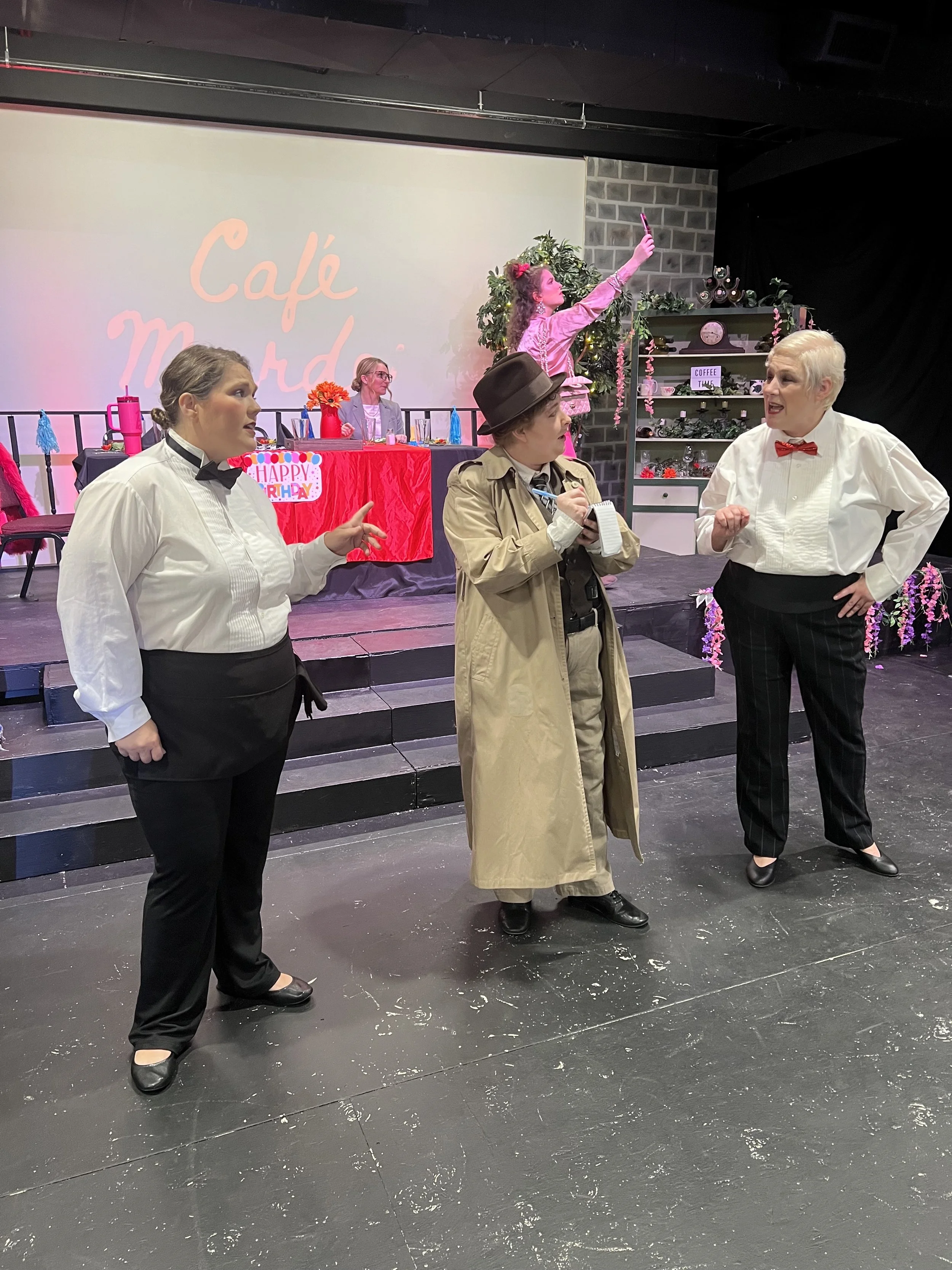 Stage scene from a play with four actors, one woman taking a selfie, a woman with a trench coat and notebook, a man in tuxedo with a bowtie, and a woman in a white shirt and black pants. In the background, a table with birthday decorations and a screen with "Café Murder" projected on it.