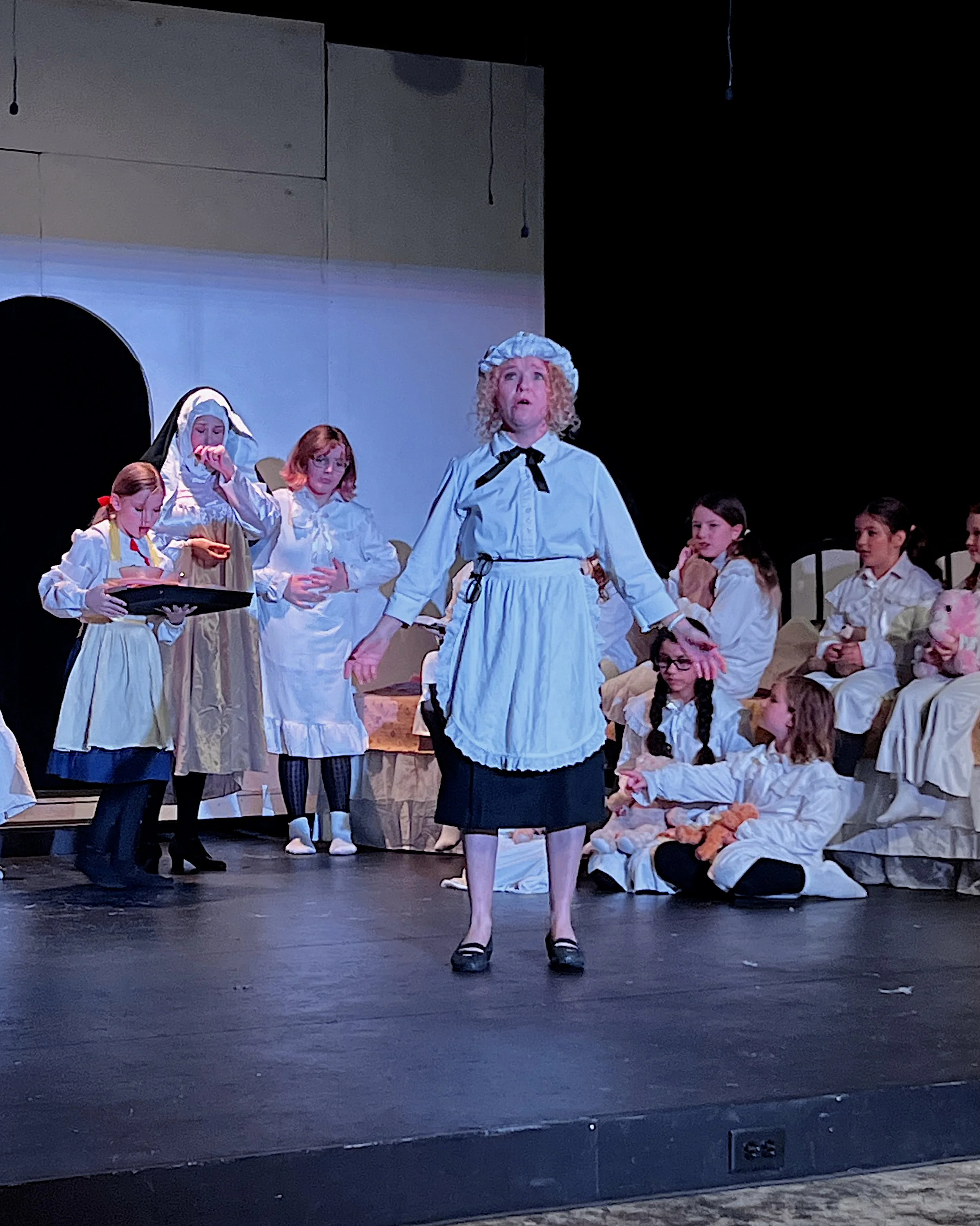 A theatrical stage performance with children dressed as characters in historical or biblical costumes. One girl in the center appears to be singing or speaking passionately, surrounded by other children seated or standing, some holding toys or props.