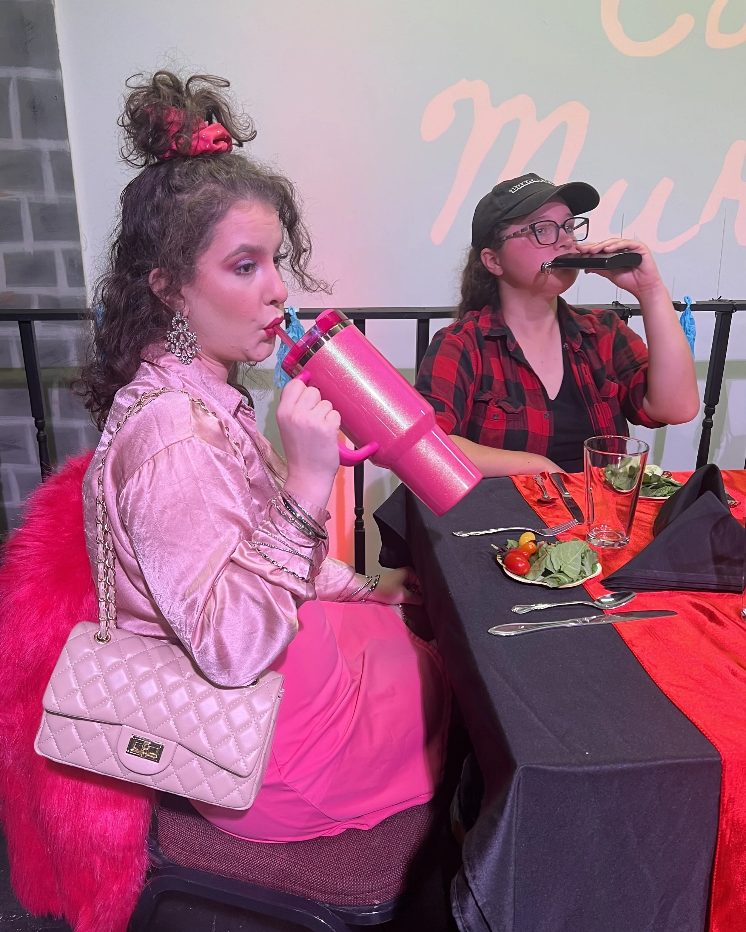 Two women sitting at a dinner table, one with curly hair in a pink dress sipping from a pink tumbler, the other with glasses and a cap playing a harmonica, with a salad and utensils on the table.