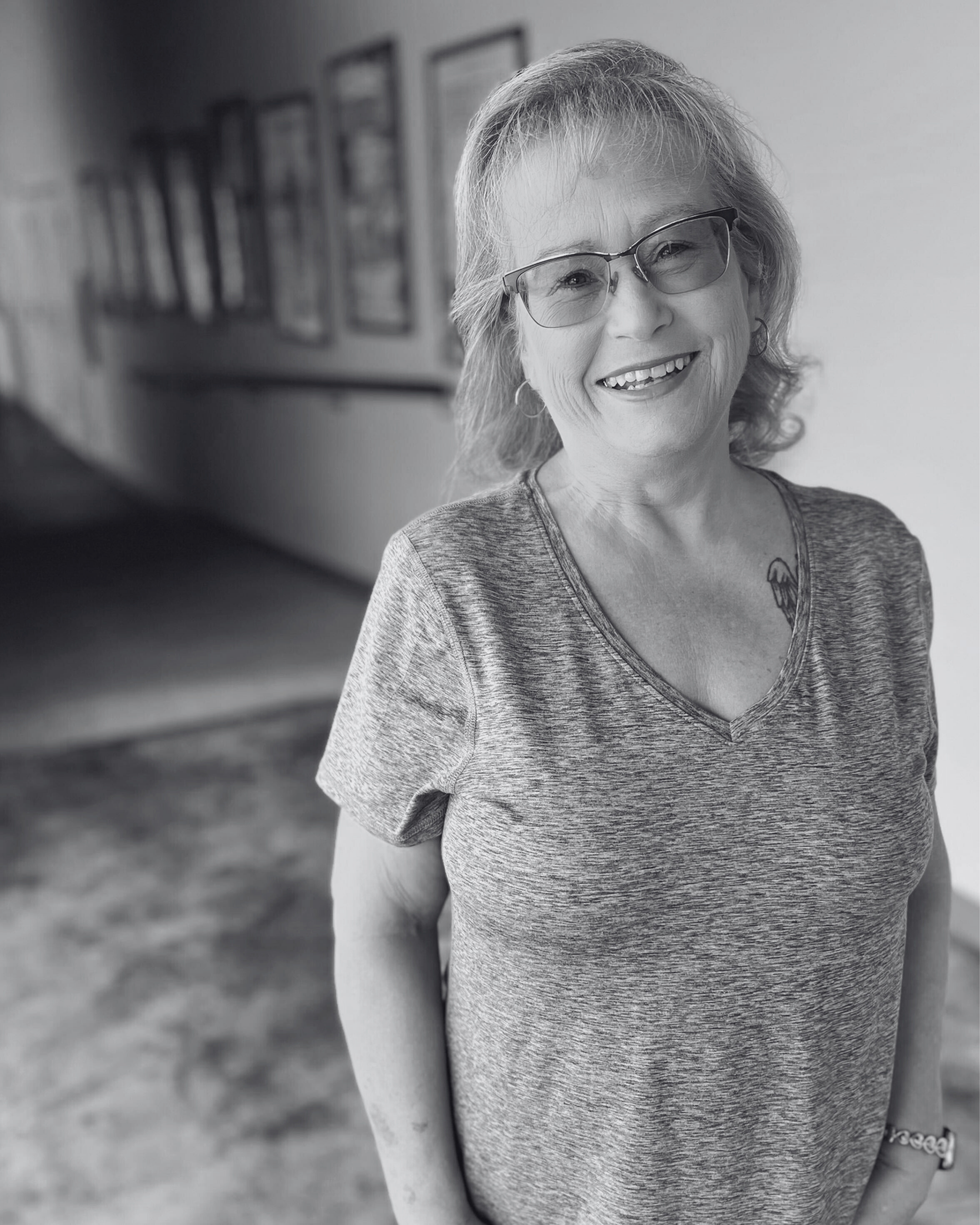 Smiling elderly woman with glasses and a tattoo on her chest, wearing a gray T-shirt and standing indoors in front of a beige wall with framed pictures.