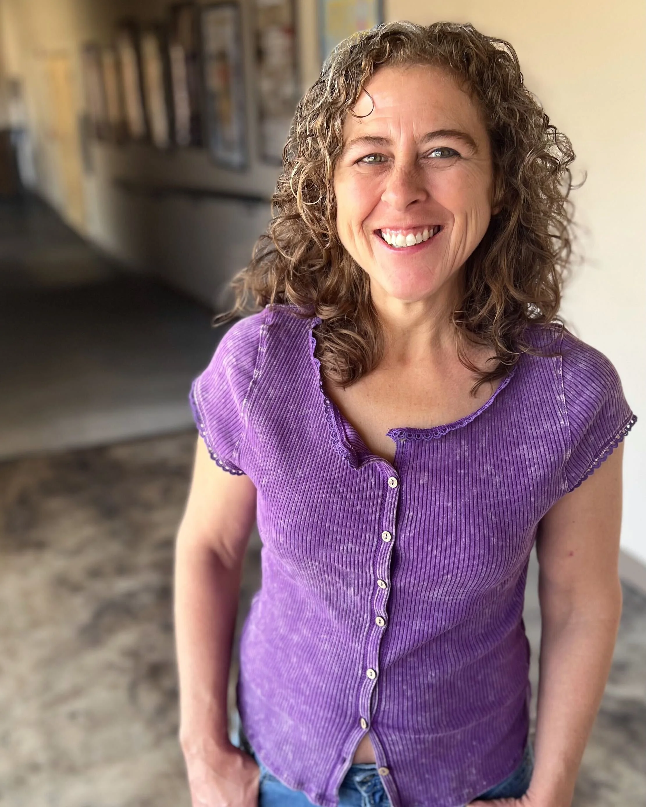 A woman with curly brown hair and a big smile, wearing a purple button-up top, standing indoors with a blurred background of art and books.