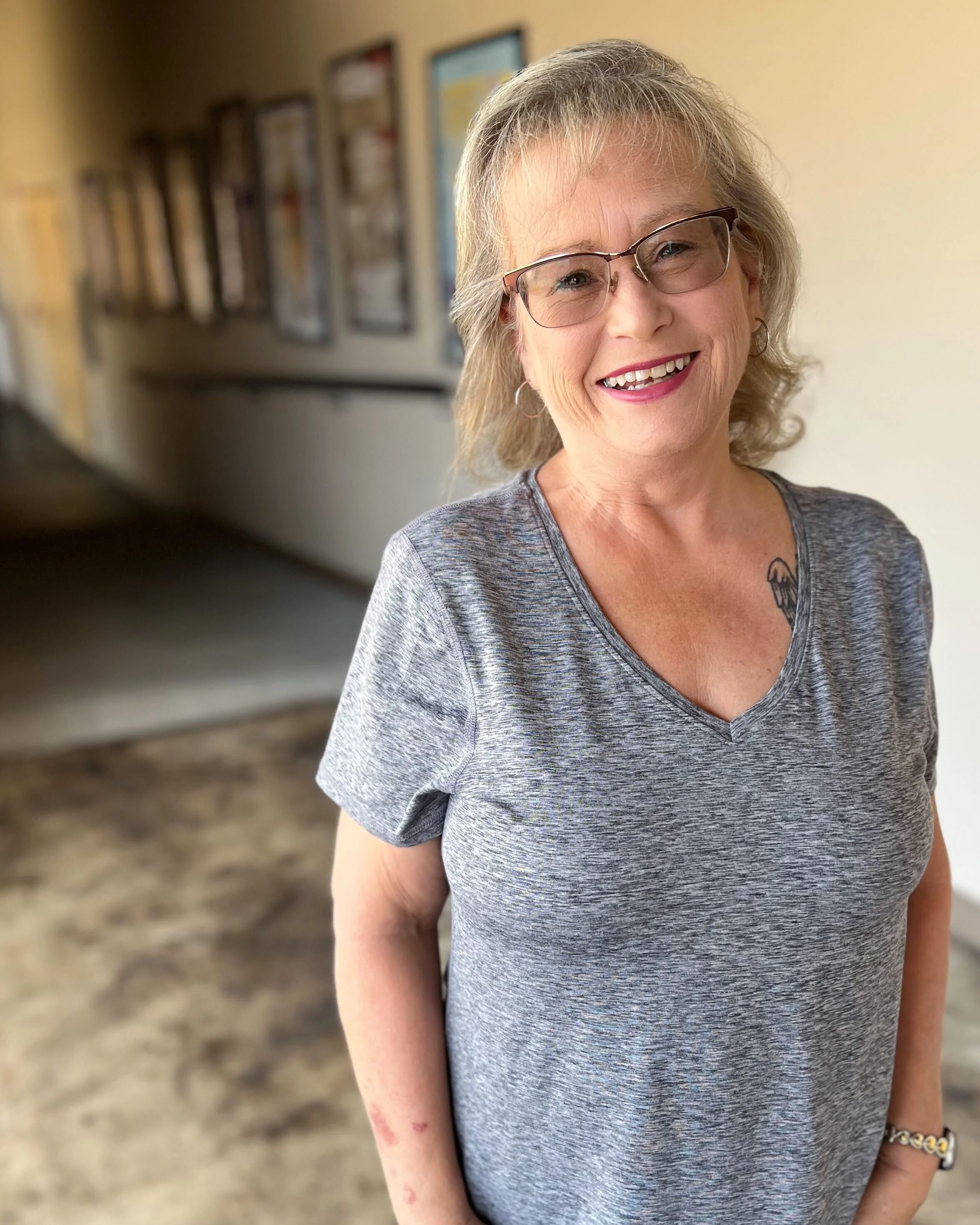 Smiling elderly woman with glasses and a tattoo on her chest, wearing a gray T-shirt and standing indoors in front of a beige wall with framed pictures.