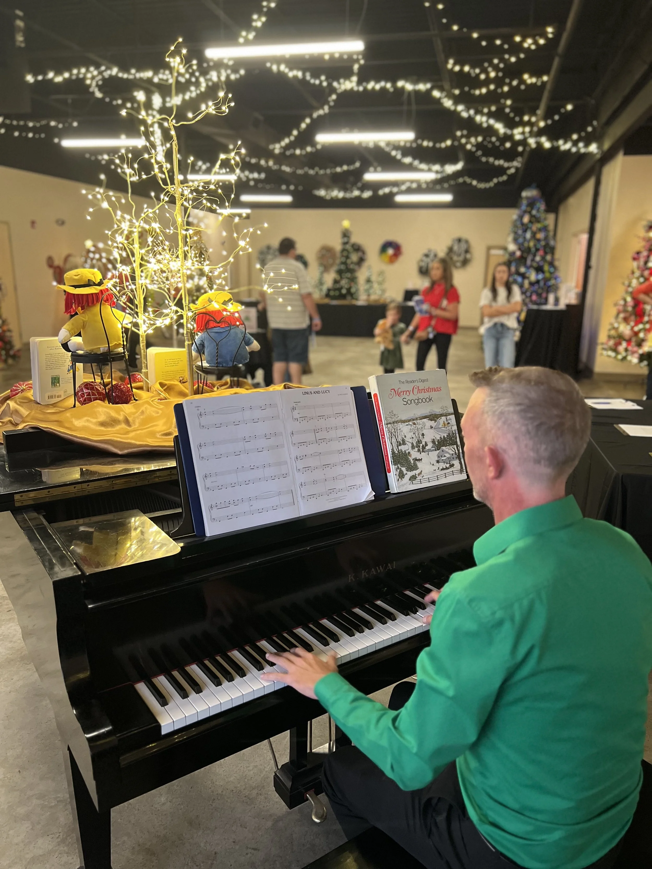 Man playing black grand piano decorated with Christmas-themed plush toys and illuminated wire trees, in a room decorated with Christmas trees and hanging fairy lights, with people in the background during a holiday event.