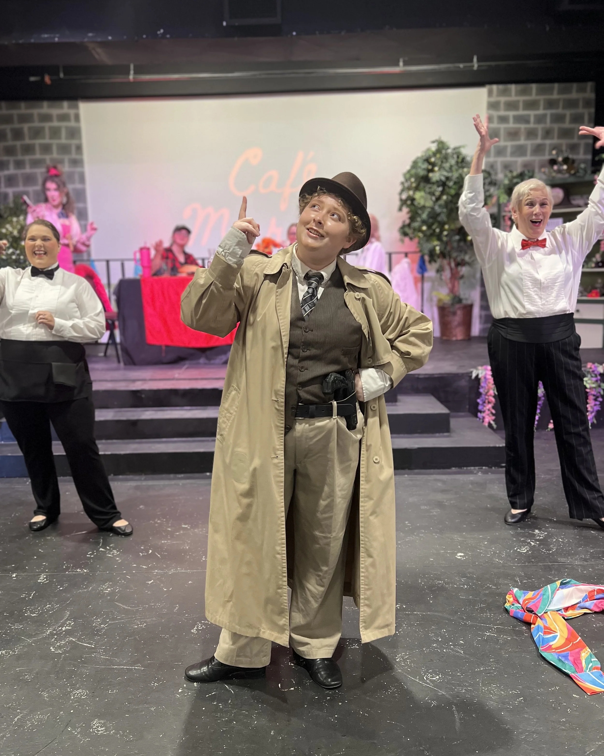Actors performing on stage in a production, with one dressed as a detective or police officer in the foreground, and others behind with joyful expressions, set with colorful props and a backdrop reading 'Café.'