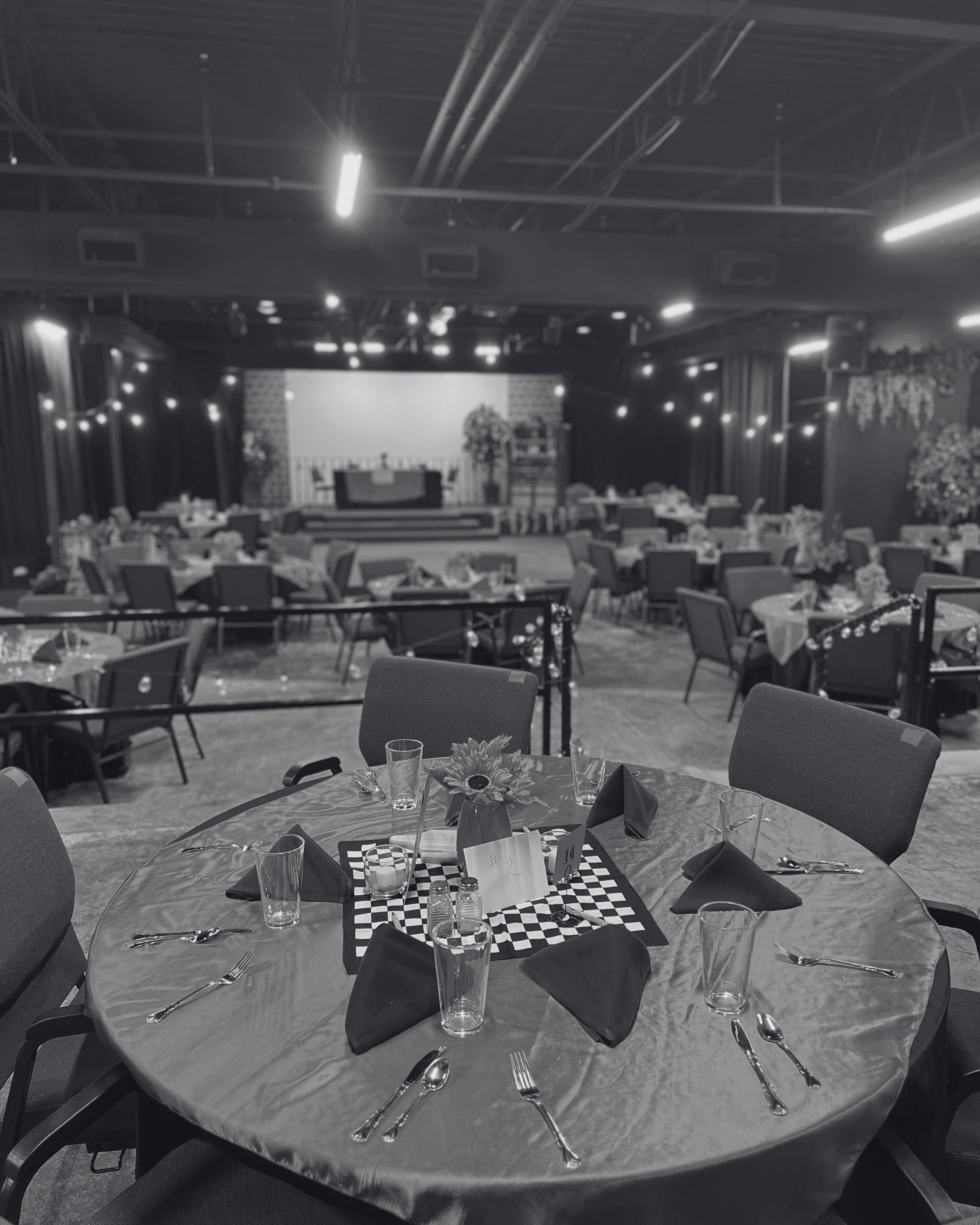 Indoor event space with tables set for a meal, a stage at the front, and string lights hanging from the ceiling.